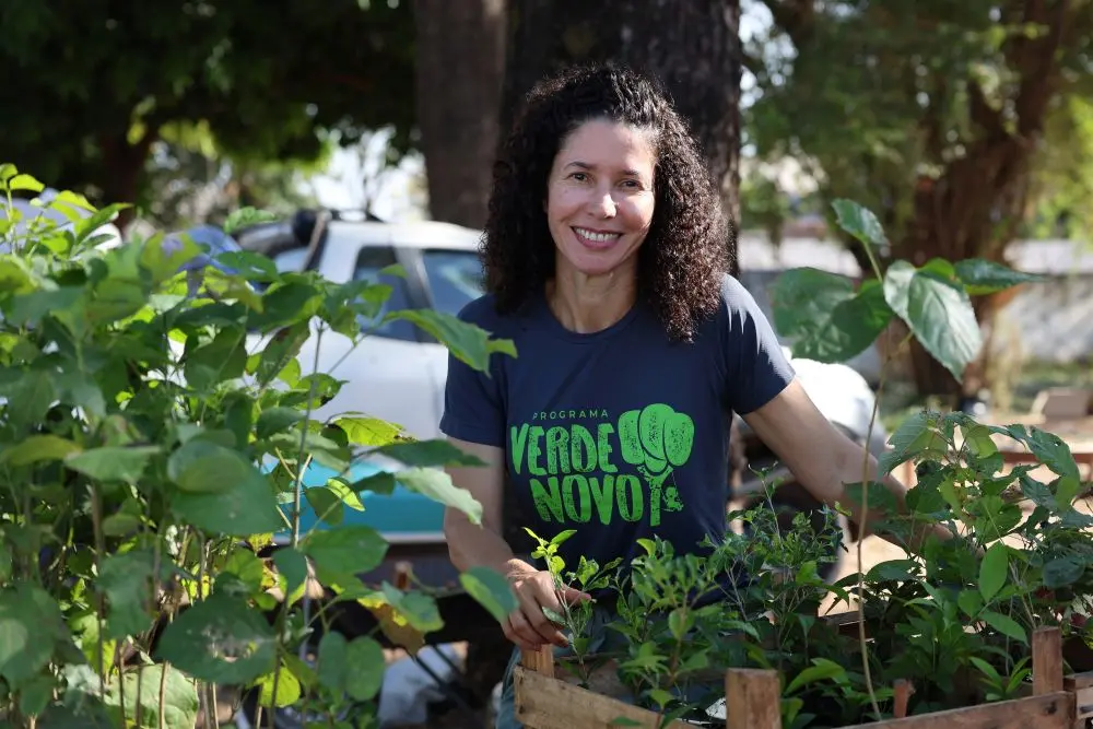 Foto horizontal que mostra a engenheira florestal do programa Verde Novo, Rosiani Carnaíba, agachada ao solo, em meio às mudas de plantas, sorrindo para a foto. Ela é uma mulher branca, de cabelos cacheados e castanhos, usando camiseta cinza com logomarca do Verde Novo. Ao fundo, árvores e um carro.