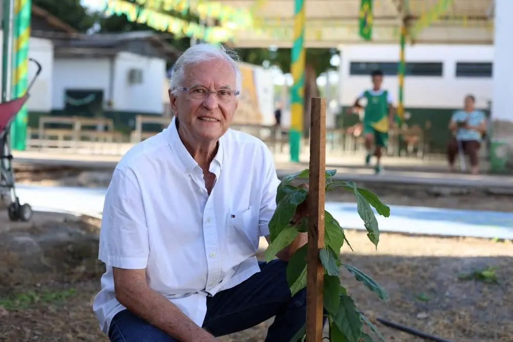 Foto horizontal que mostra o secretário de Planejamento Urbano de Cuiabá, José Afonso Portocarrero, agachado ao solo, ao lado de uma muda de árvores plantada no pátio da escola municipal Dom Bosco. Ele é um senhor branco, de cabelos brancos, usando camisa branca, calça jeans e óculos de grau. 