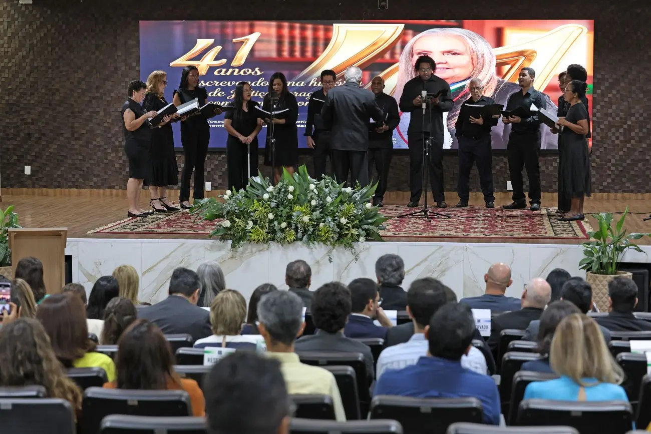Foto horizontal em plano aberto que mostra o Coral do TJMT se apresentando no palco, com todos os integrantes vestidos de preto. A plateia está lotada. O telão exibe uma foto da desembargadora Maria Erotides Kneip.