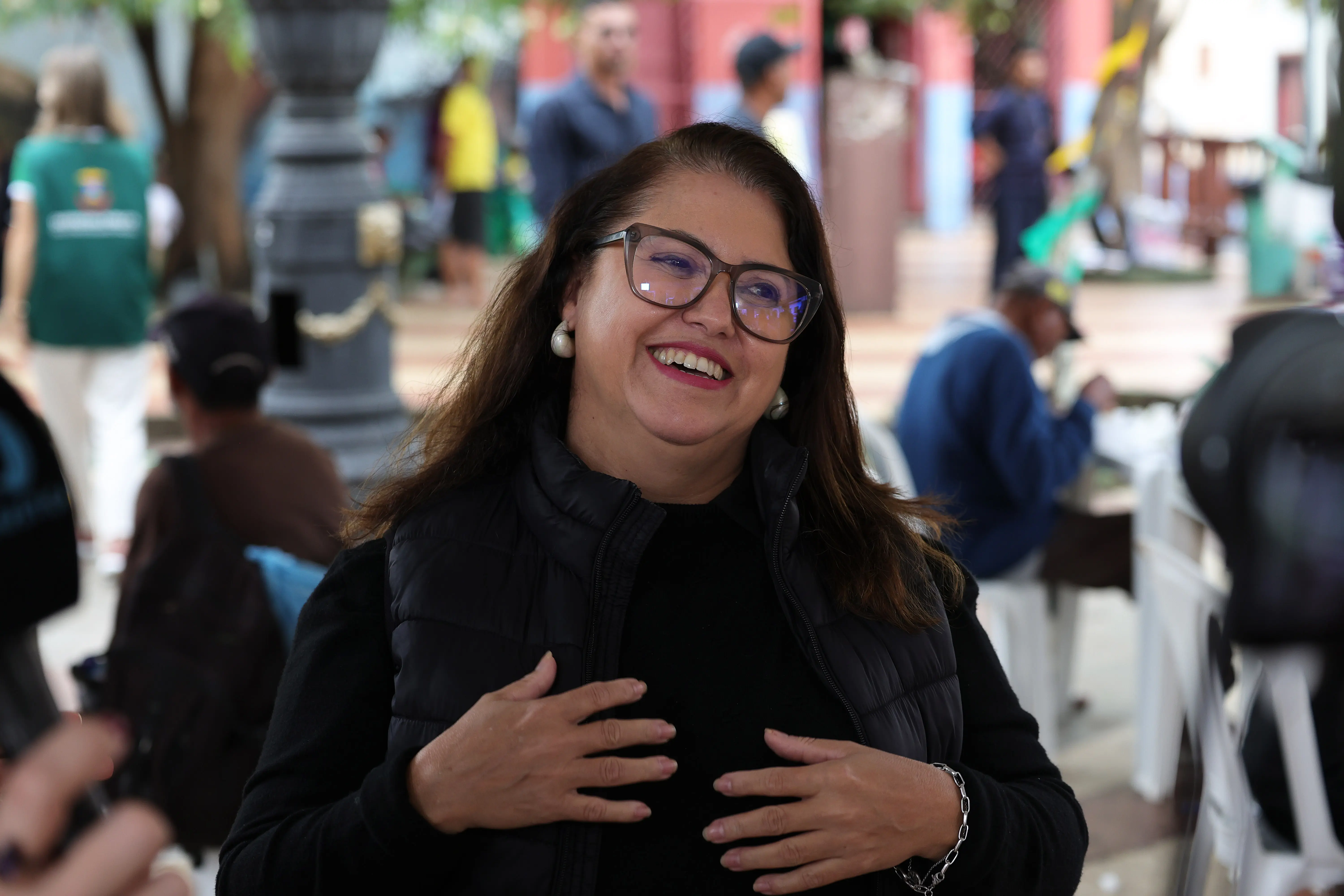 Mulher com cabelos longos e castanhos escuros. Ela está sorrindo e olhando para cima e para a esquerda, com as mãos apoiadas no peito. Ela veste um colete preto sobre uma blusa preta e usa óculos de grau. O fundo mostra uma praça pública com pessoas e vegetação embaçadas.