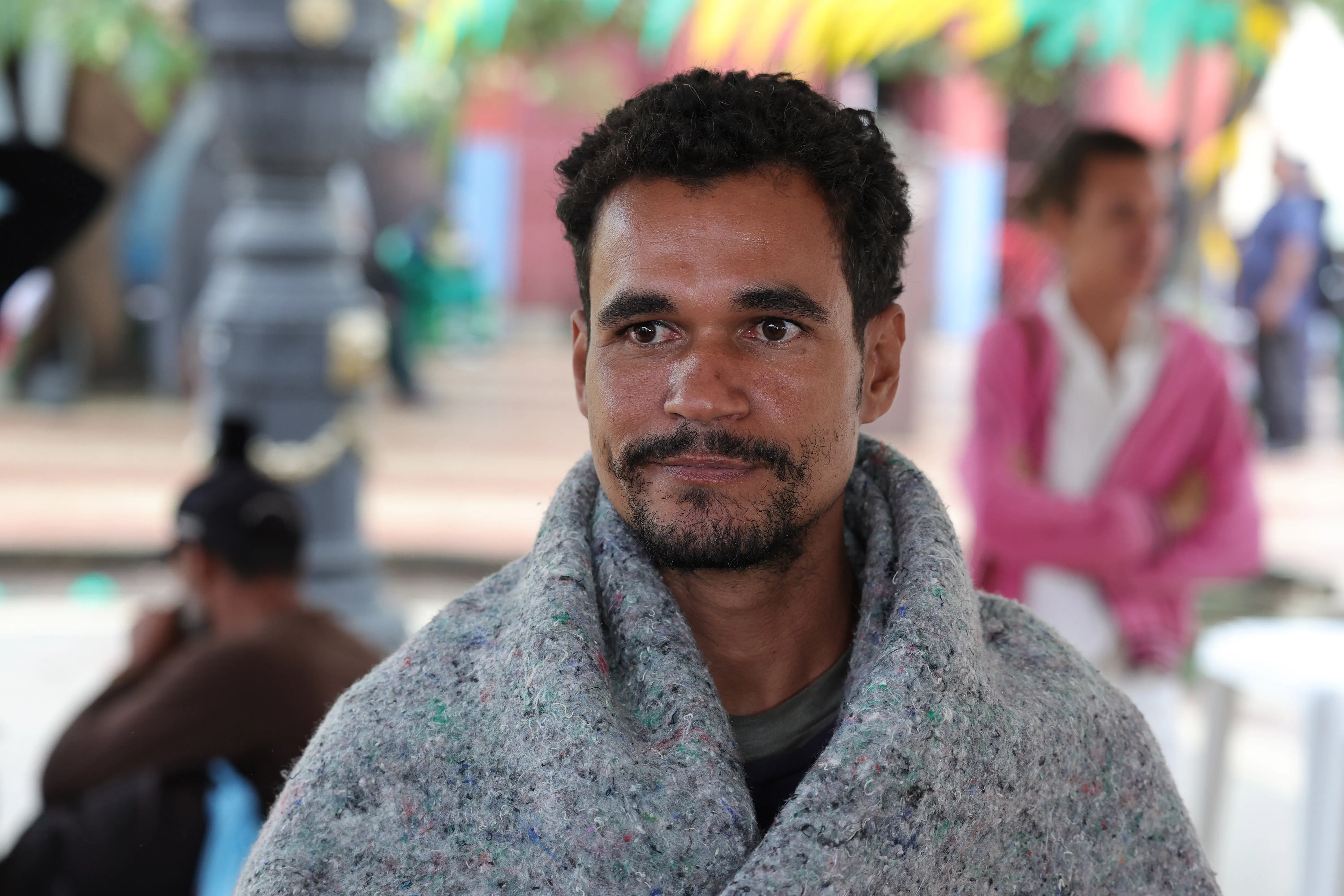 homem com cabelos curtos e cacheados pretos e barba rala. Ele está envolto em um cobertor cinza texturizado sobre os ombros e olha ligeiramente para a esquerda. Ele usa uma camiseta escura por baixo do cobertor. O fundo embaçado mostra uma praça pública com pessoas e uma estrutura de luz.