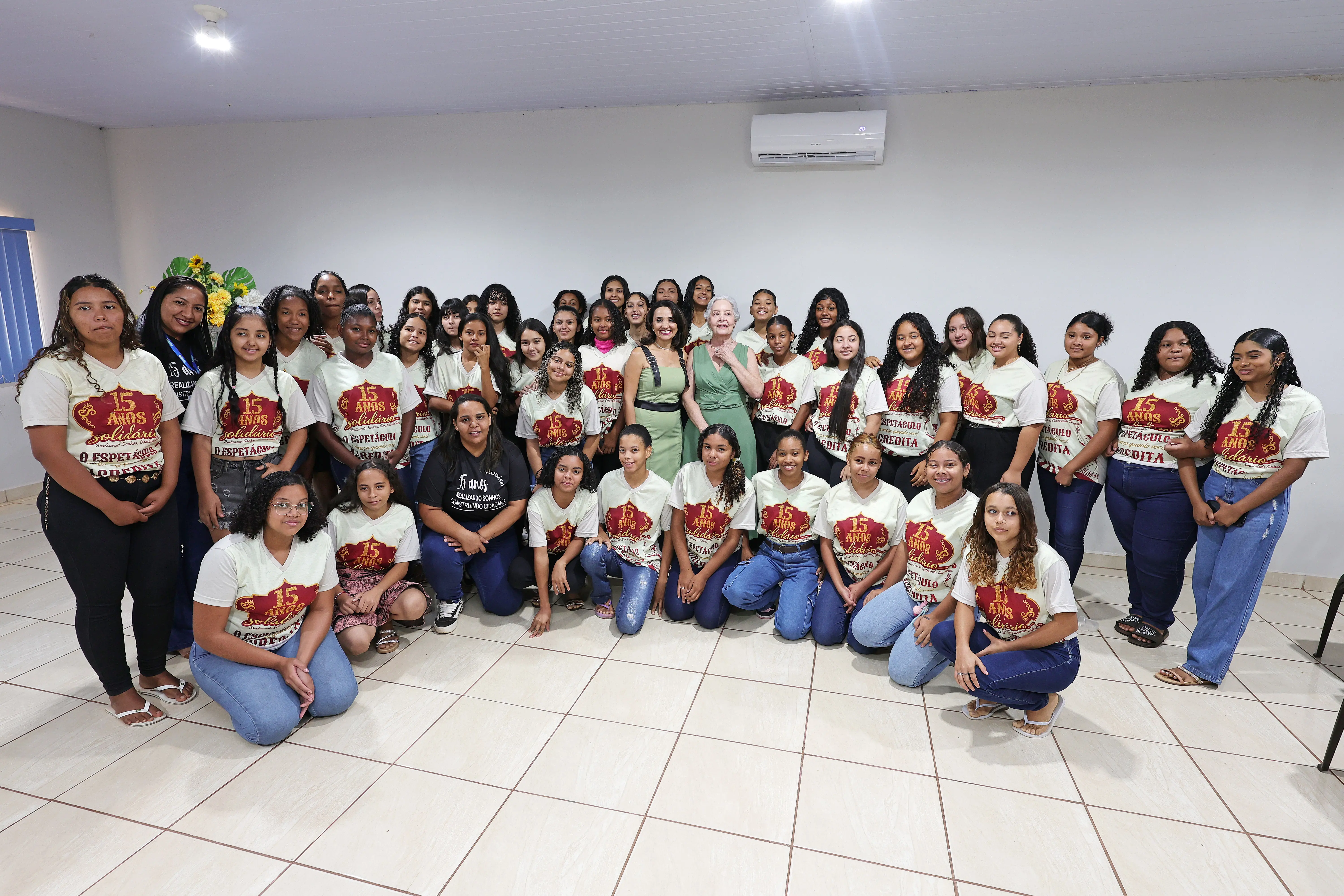 Grande grupo de jovens posa para foto em um ambiente interno. A maioria veste camisetas beges com estampa vermelha. Duas mulheres, uma de terno preto e outra de vestido verde, estão no centro.