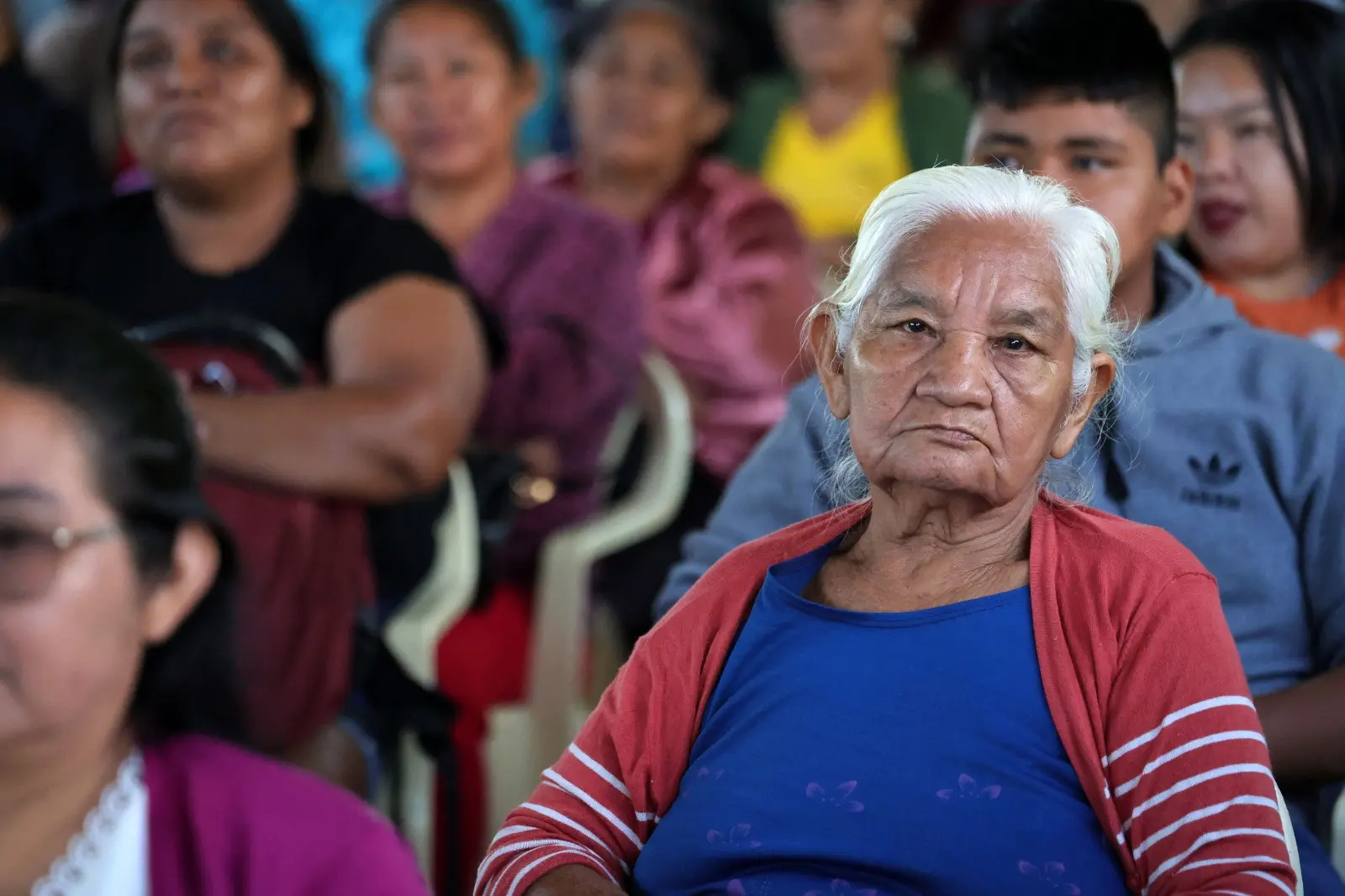 Mulheres indígenas e outras pessoas estão sentadas em cadeiras plásticas sob uma tenda. Ao fundo, um painel com os logos do Poder Judiciário de MT e a frase "JUSTIÇA SEM FRONTEIRAS".