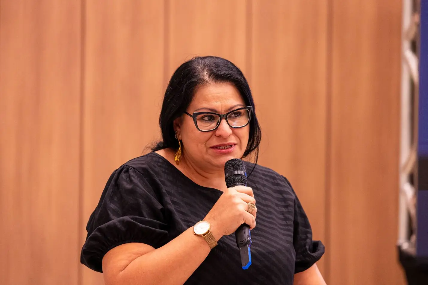 Foto horizontal que mostra a agente de captação do MT Hemocentro, Magda Matos, falando ao microfone. Ela é uma mulher de pele parda, com cabelos longos, lisos e pretos, presos para trás, olhos castanhos escuros, usando óculos de grau e blusa preta. 