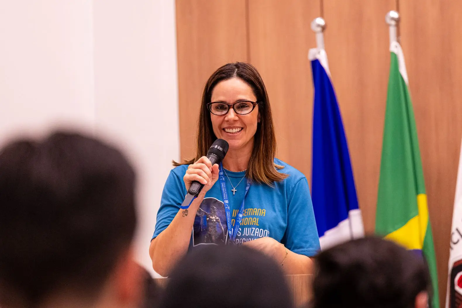 Foto horizontal que mostra a diretora do Daje, Shusiene Machado, falando ao microfone, em pé, em um auditório. Ela é uma mulher branca, de cabelos lisos, castanhos na altura dos ombros, usando camiseta azul com arte da Semana dos Juizados Especiais.