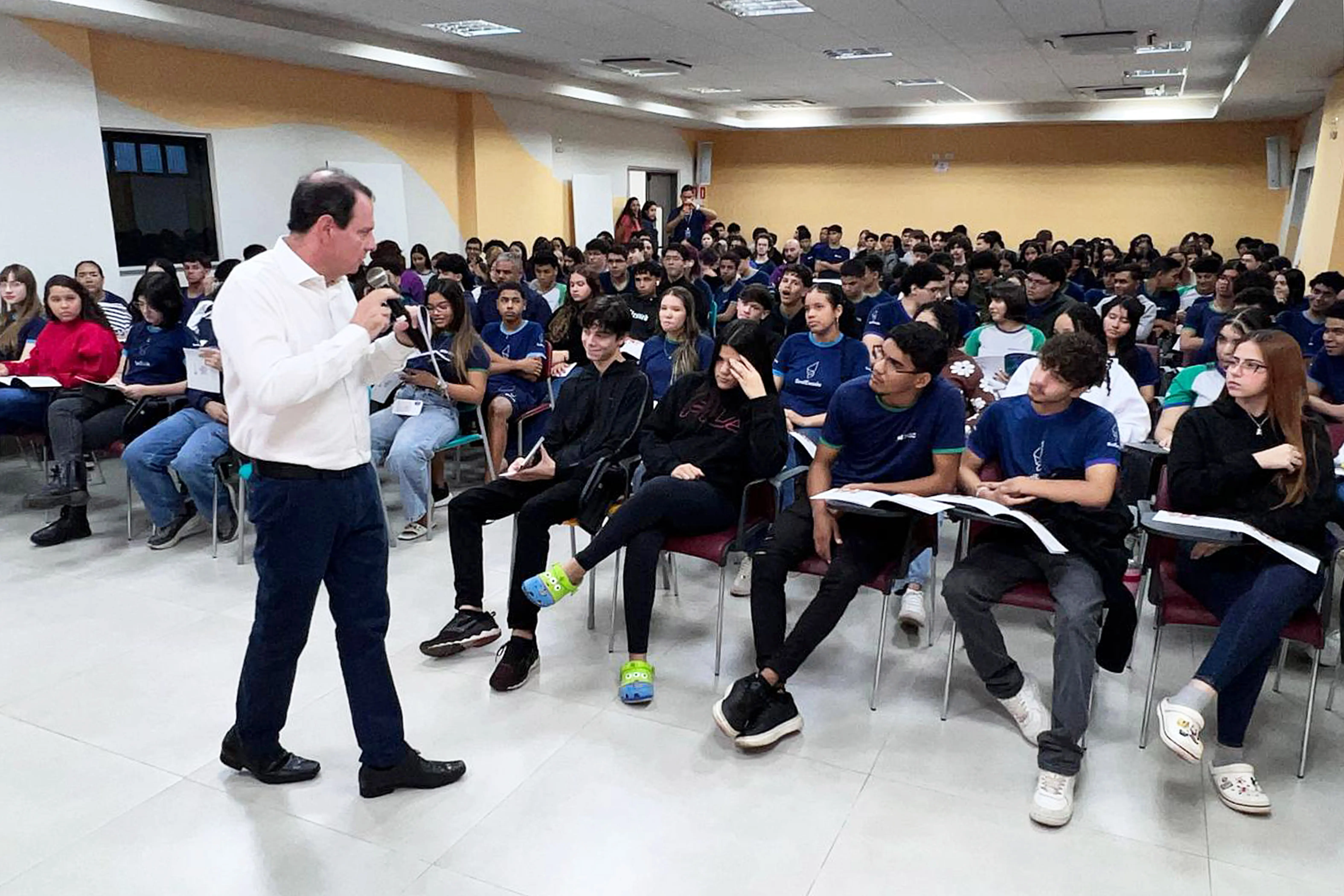 Imagem de ângulo médio mostrando um homem de camisa branca e calça azul falando ao microfone para um grande público sentado em cadeiras de auditório. A plateia é jovem, composta por estudantes em sua maioria com uniformes azuis e pretos.