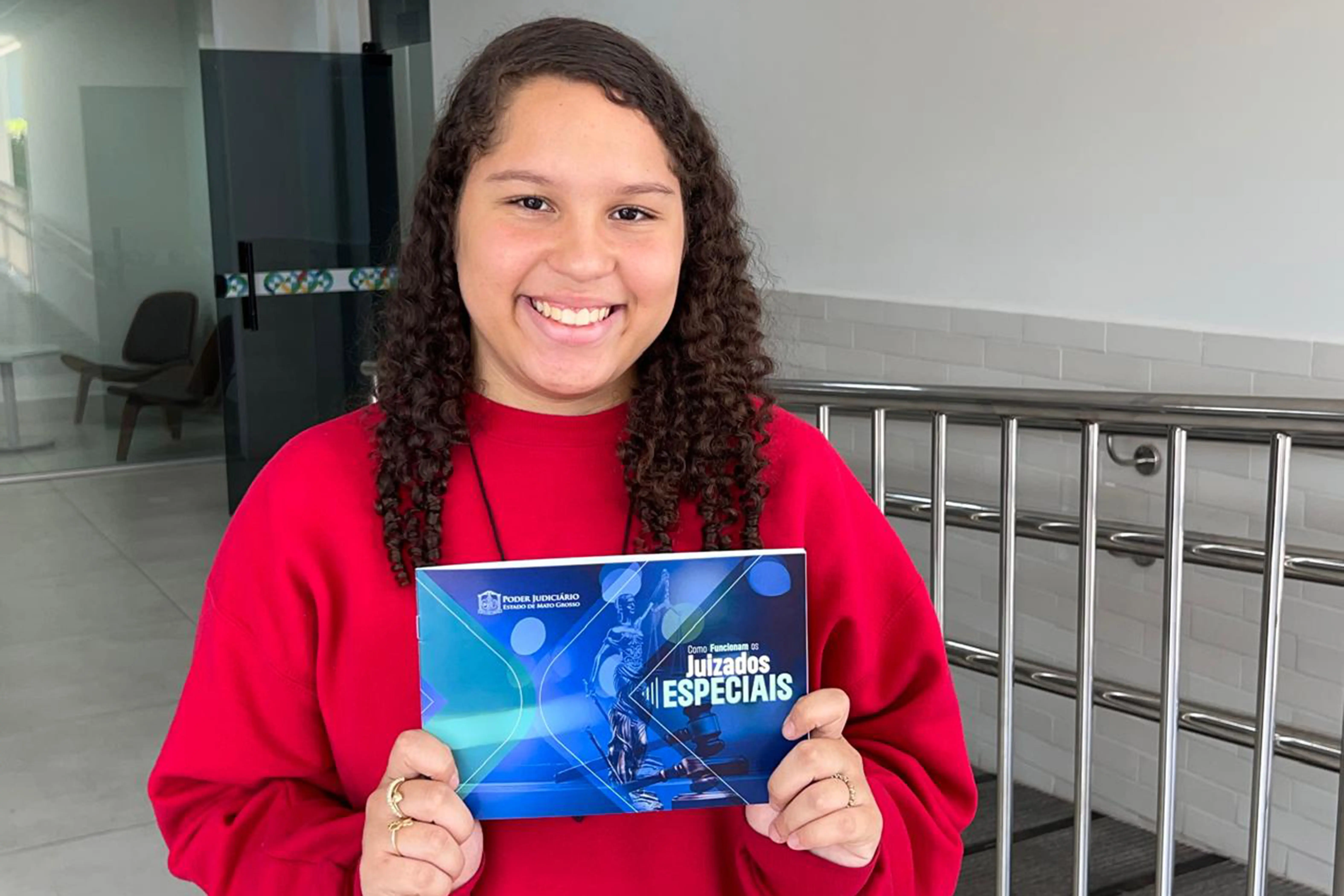Menina adolescente sorridente, com cabelos cacheados, veste um casaco vermelho vivo e segura um folheto do "Poder Judiciário de Mato Grosso" com o título "Como Funcionam os Juizados Especiais". Ela está em pé em um ambiente de corredor com corrimãos de metal.