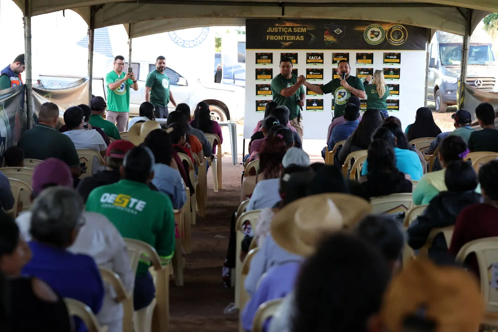 Sob uma grande tenda, o público assiste em cadeiras plásticas a dois homens com camisetas verdes que se cumprimentam no palco. Ao fundo, há um painel informativo e veículos estacionados.