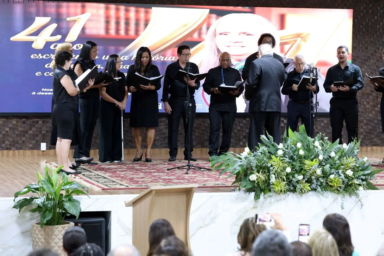 Foto horizontal em plano aberto que mostra o Coral do TJMT se apresentando no palco, com todos os integrantes vestidos de preto. O telão ao fundo exibe uma foto da desembargadora Maria Erotides Kneip.