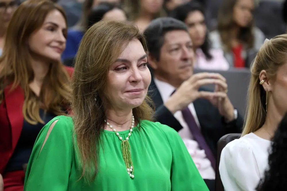 Foto horizontal que mostra Cassyra Vuolo sentada na plateia do auditório do TJMT, do busto pra cima, com olhos marejados. Ela é uma mulher branca, de longos cabelos lisos e castanhos, usando vestido verde e colar de pérolas.