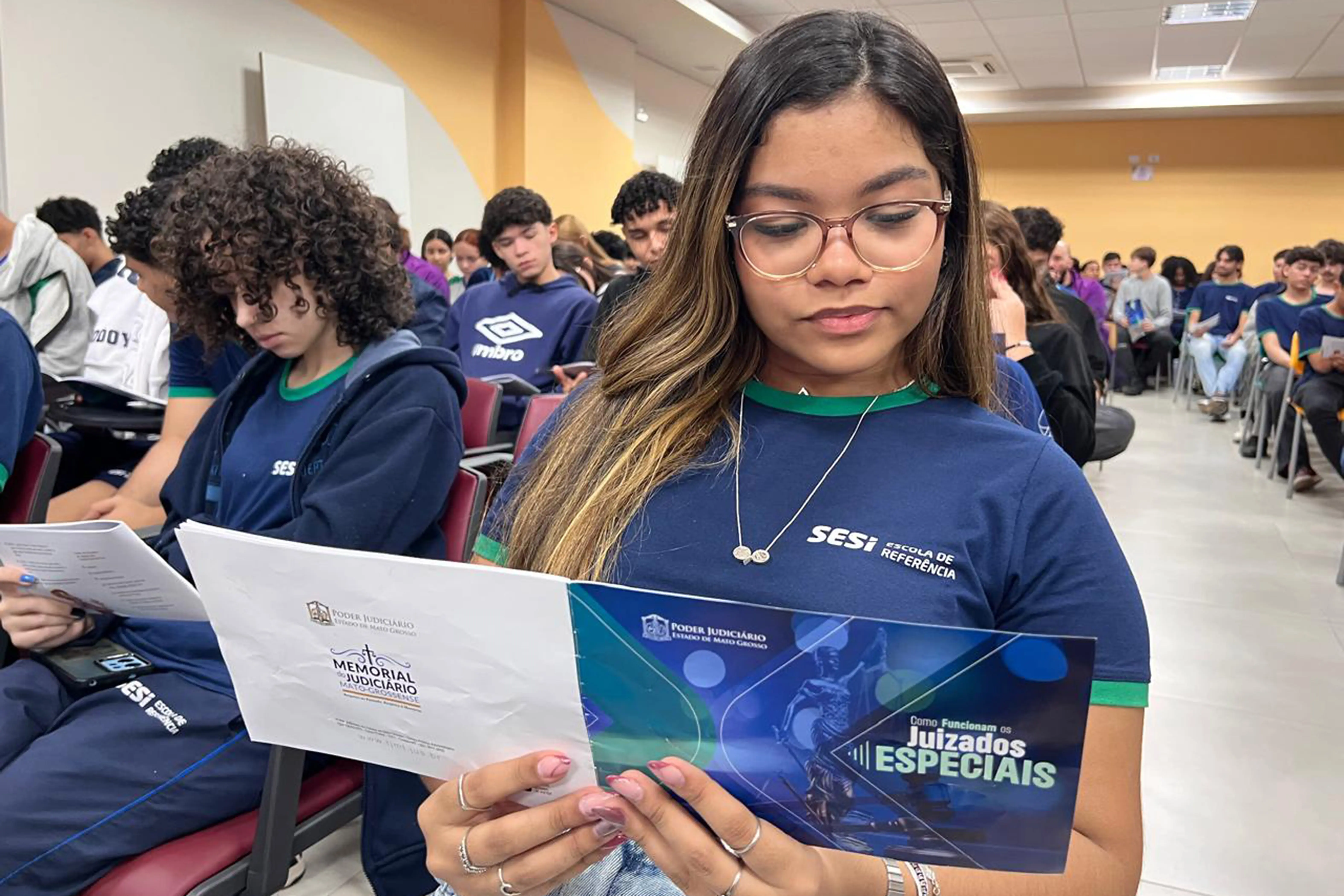 Foto de uma adolescente de óculos sentada, com camiseta azul marinho do SESI e detalhes verdes, segurando um folheto intitulado "Como Funcionam os Juizados Especiais". Outros estudantes uniformizados estão sentados em cadeiras de auditório ao fundo.