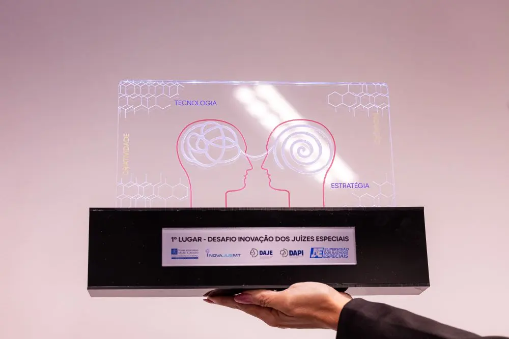 Foto horizontal de uma mão segurando a placa do Prêmio Desafio Inovação dos Juizados Especiais. A placa é de acrílico transparente sobre uma base preta. Na placa há a imagem de duas cabeças com os cérebros representados por fios que se conectam.
