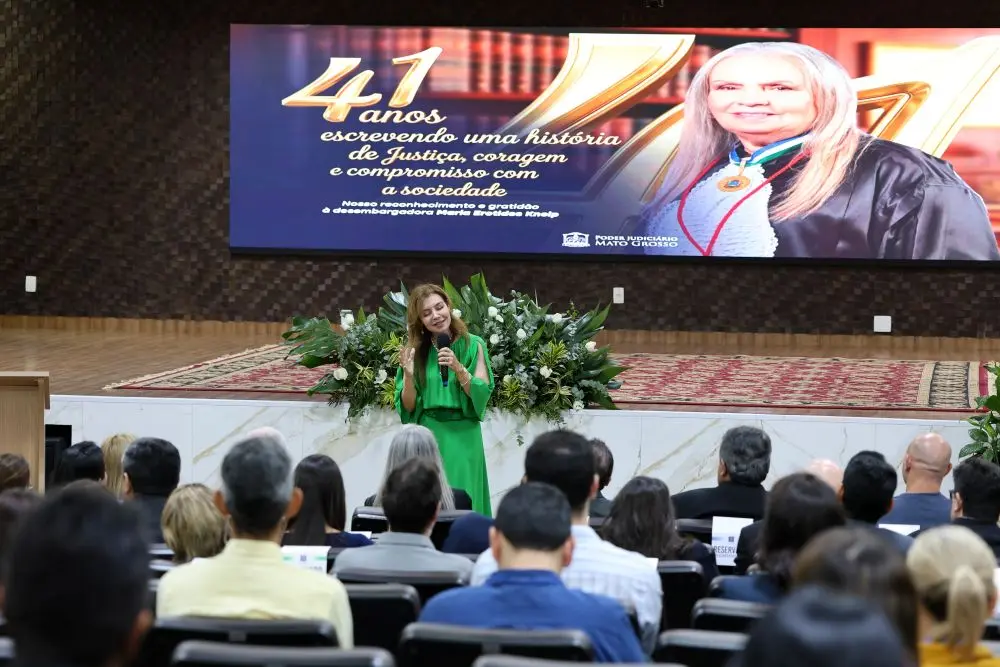 Foto horizontal em plano aberto que mostra o auditório do TJMT lotado de pessoas na plateia, assistindo Cassyra Vuolo, que está a frente, falando ao microfone. Ela é uma mulher branca, de cabelos longos e castanhos, usando vestido verde. Ao fundo, telão exibe foto da desembargadora Maria Erotides.