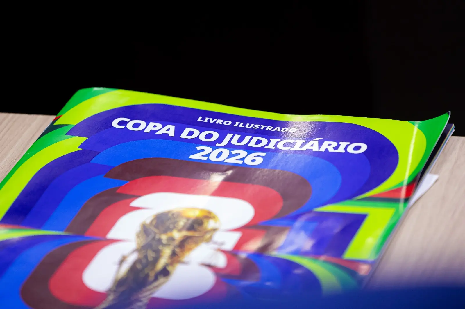 Detalhe do álbum ilustrado Copa do Judiciário 2026 sobre uma mesa. A capa colorida traz elementos gráficos inspirados nos álbuns de figurinhas das Copas do Mundo e exibe o título da publicação em destaque.