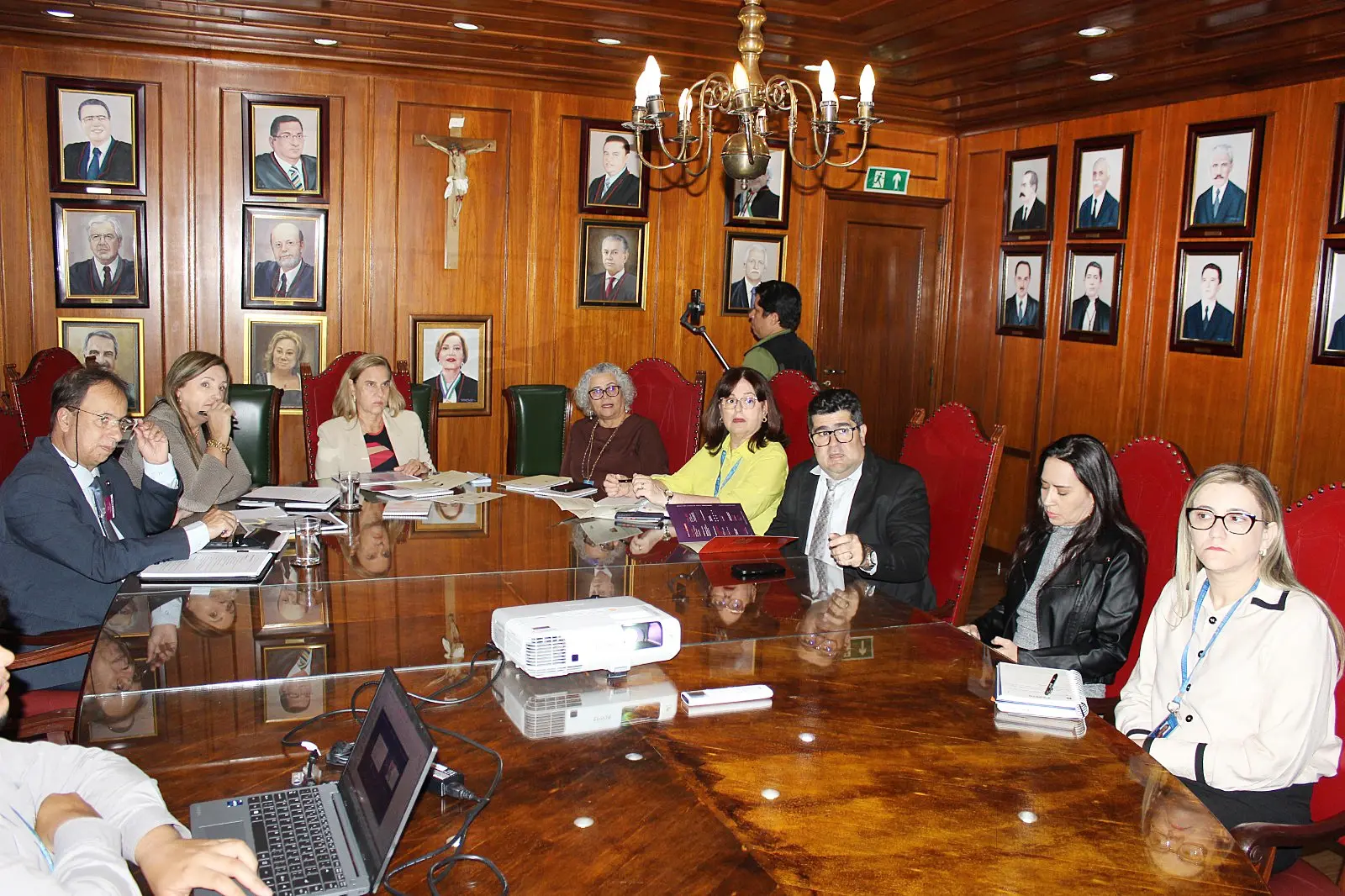 A imagem mostra a sala de reunião do Espaço Memória, no TJMT. Nove pessoas estão sentadas em volta da mesa. Olhando atentamente para o telão que não aparece na foto. 