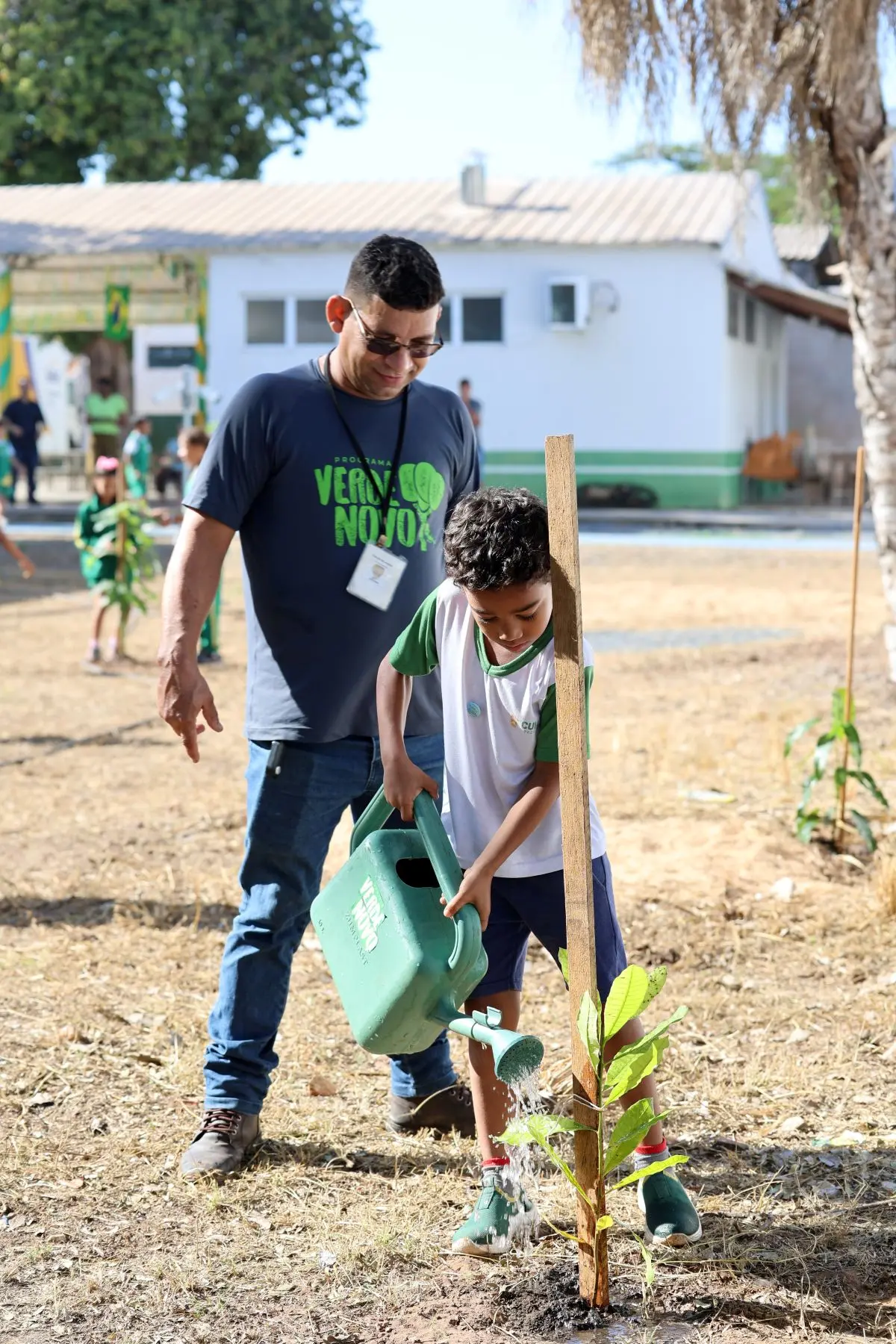 Foto vertical que mostra um menino com uniforme verde e branco regando uma muda de caju plantada no pátio da escola municipal Dom Bosco. O regador é verde e tem a logomarca do programa Verde Novo. Ao lado dele, um servidor usando camiseta cinza com a logomarca do programa Verde Novo, observa e sorri
