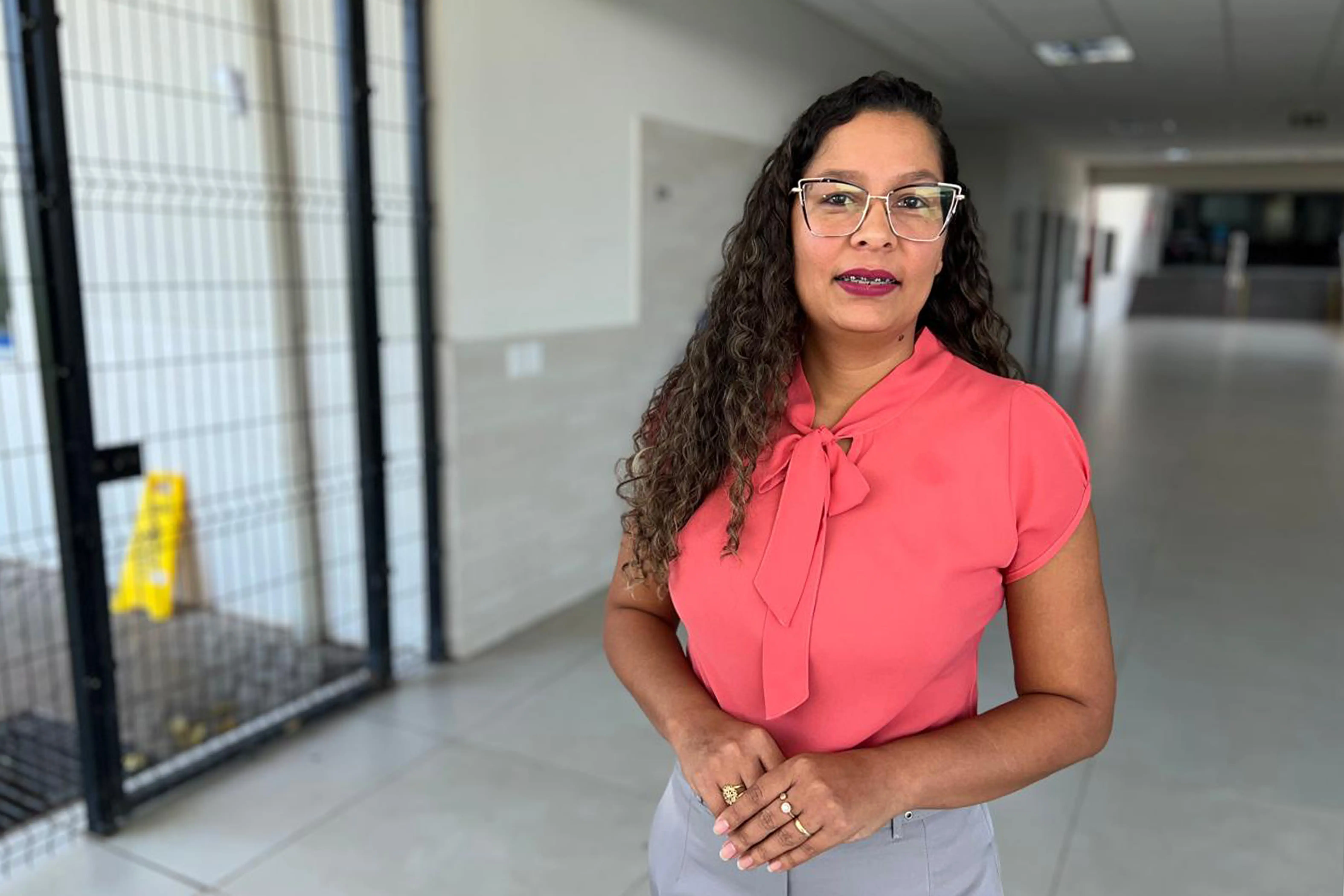 Retrato ede uma mulher de óculos, com cabelos cacheados e longos, vestindo uma blusa coral com laço no pescoço e calça cinza. Ela sorri para a câmera com as mãos entrelaçadas. O fundo é um corredor claro com uma grade preta e uma placa amarela de alerta.