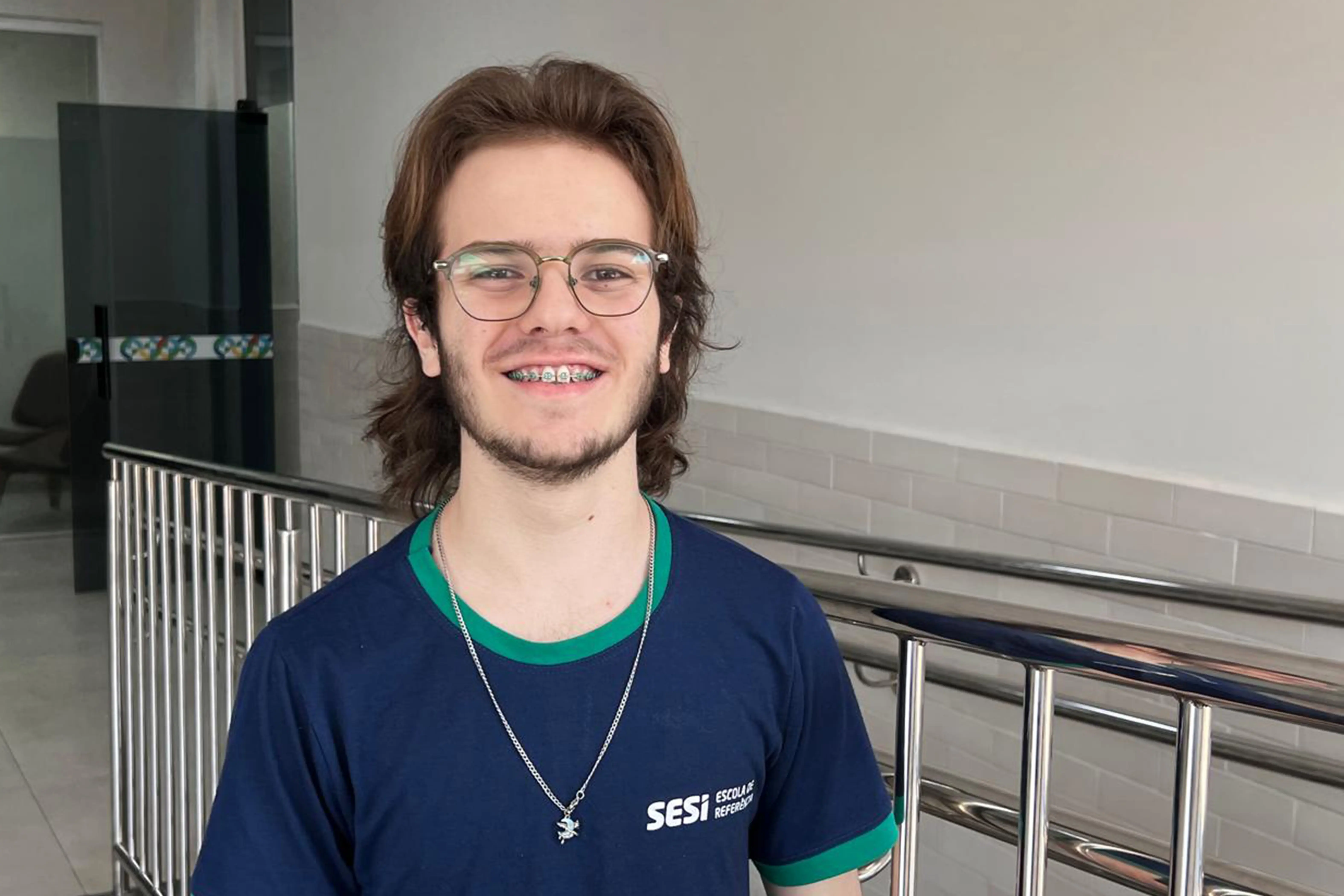 Retrato de um adolescente com óculos e aparelho ortodôntico, sorrindo para a câmera. Ele veste uma camiseta azul marinho do SESI, com gola e mangas verdes, e um colar de corrente com um pingente. Ao fundo, um corredor com corrimãos de metal.