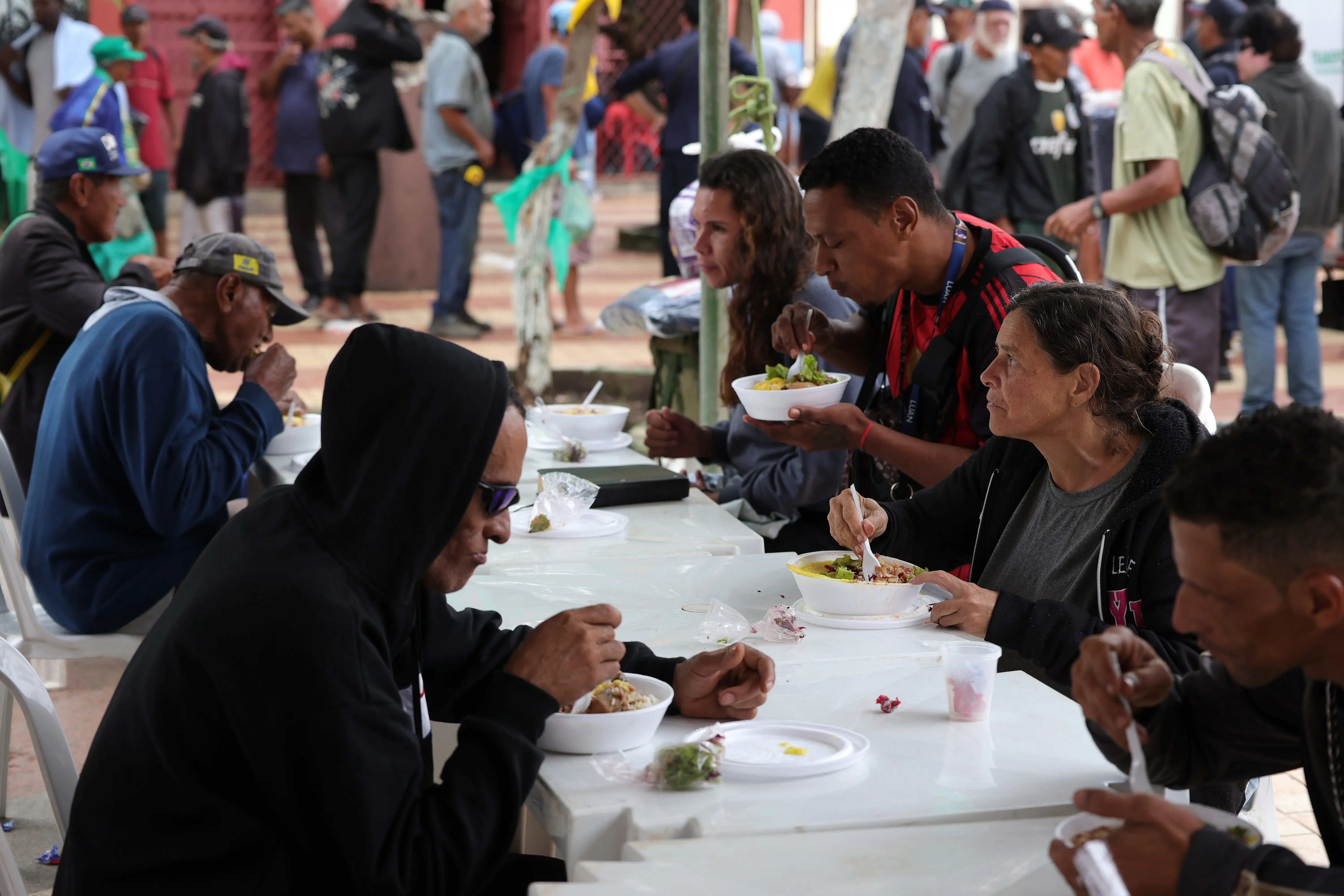 Grupo de pessoas sentadas em mesas de plástico brancas em uma praça pública. Várias pessoas estão comendo refeições em tigelas brancas. Elas estão vestidas com roupas casuais, incluindo casacos e bonés. O ambiente é movimentado, com outras pessoas em pé ao fundo e vegetação.
