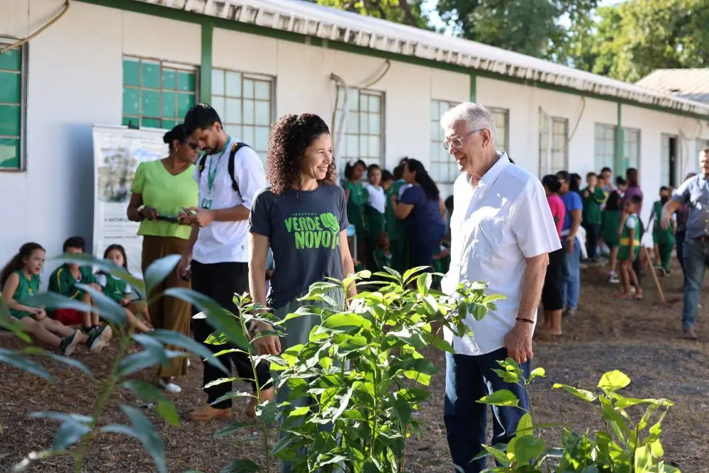Foto horizontal em plano aberto que mostra, a engenheira florestal do Verde Novo, Rosiani Carnaíba, e o secretário de Planejamento Afonso Portocarrero, conversando no pátio da escola Dom Bosco. Na frente deles, há várias mudas de plantas. Ao fundo, movimentação de alunos e pessoas na escola.