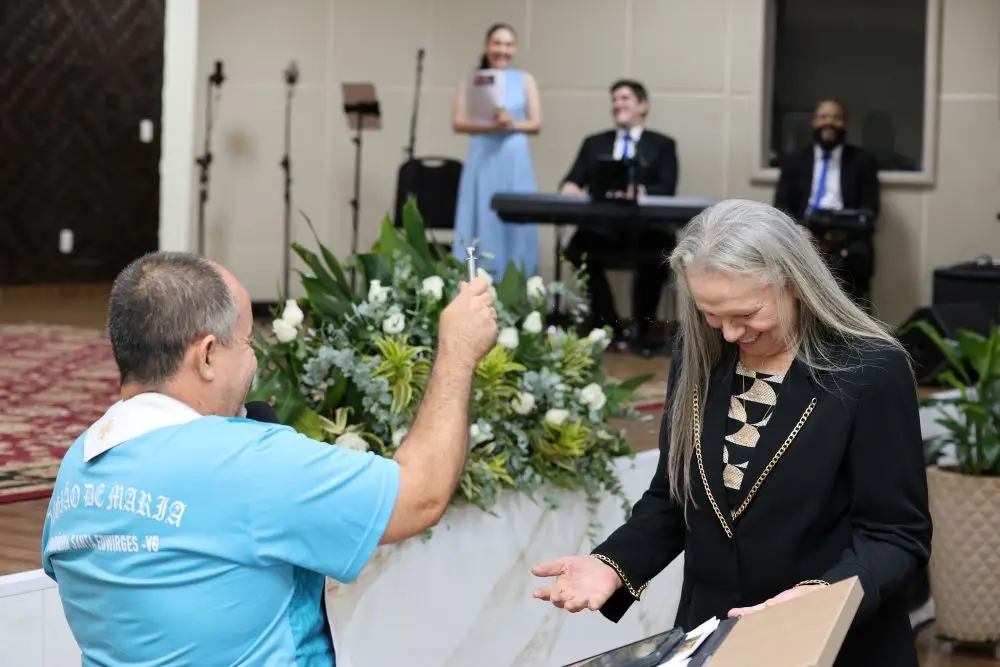 Foto horizontal que mostra o padre Divair, de costas, aspergindo água benta na desembargadora Maria Erotides Kneip, que está com as mãos em reverência e cabeça abaixada, sorrindo. Ela é uma senhora branca, de longos cabelos lisos e grisalhos, usando blazer preto com detalhes dourados na gola. 