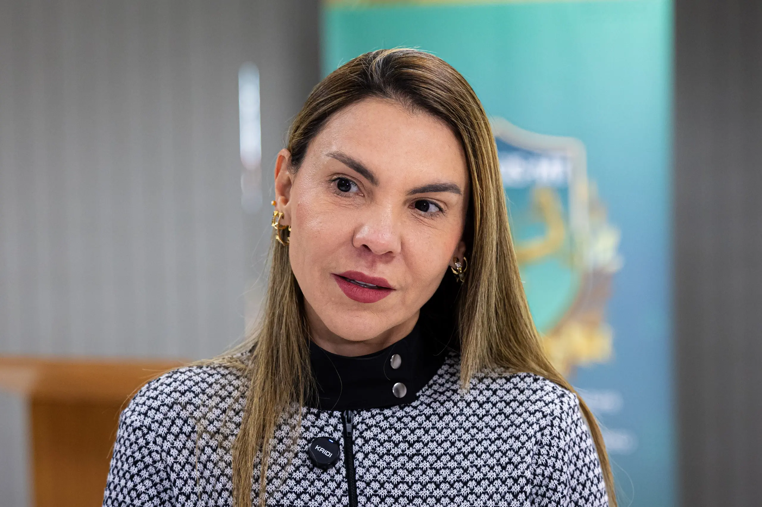 A imagem em plano médio de mulher com cabelos longos e lisos com mechas loiras, olhando ligeiramente para a direita da cena. Ela veste uma blusa com estampa geométrica quadriculada em preto e branco com gola alta preta, e um pequeno microfone de lapela está preso à roupa.