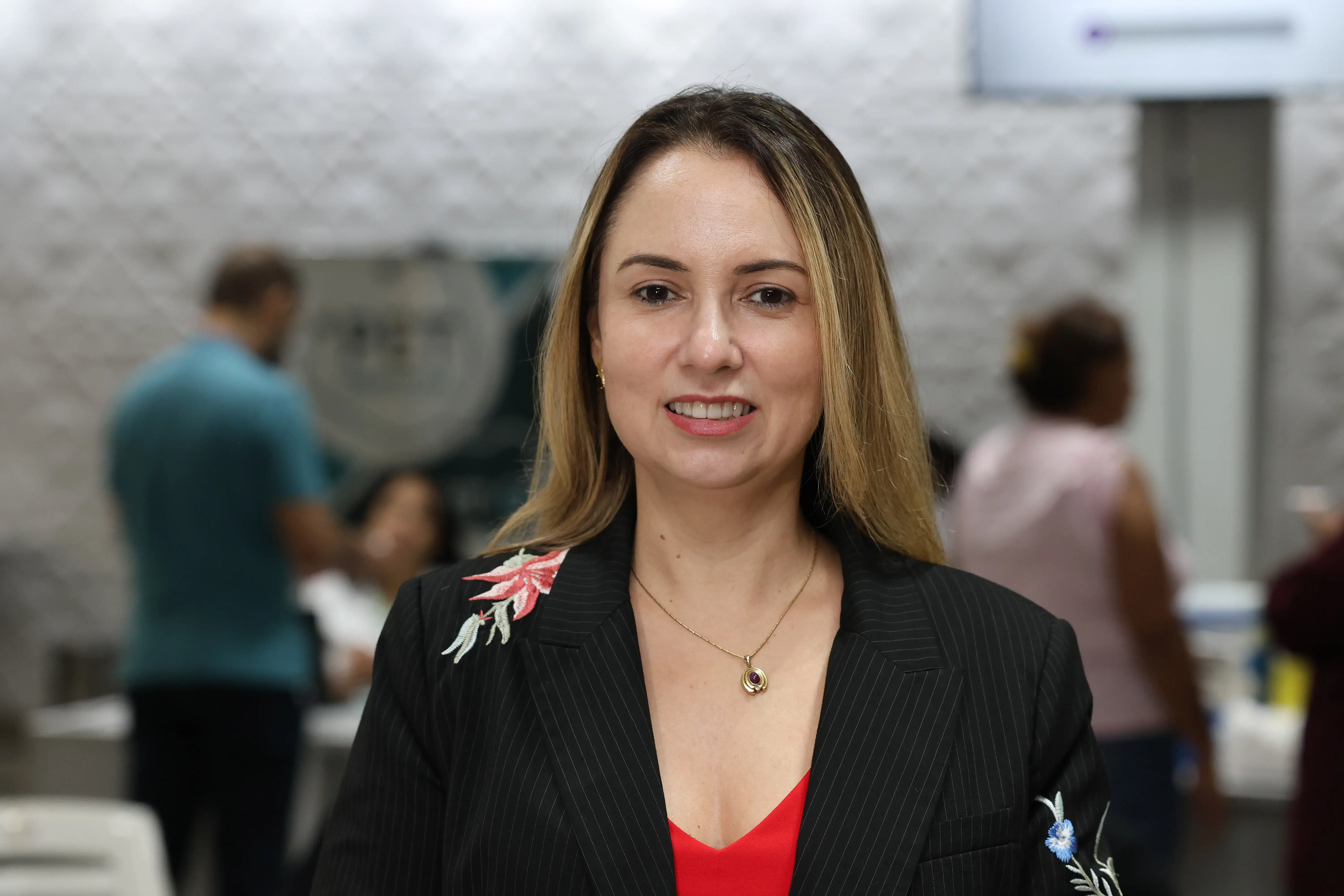 Mulher de cabelos claros sorri em primeiro plano. Usa blusa vermelha e blazer preto risca de giz com flores bordadas. Ao fundo desfocado, há pessoas em um ambiente interno.