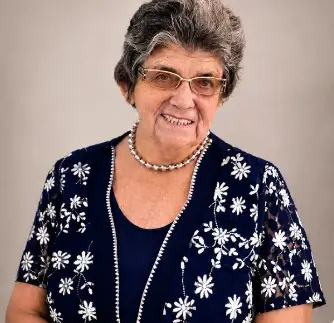 Mulher idosa sorrindo, de óculos e cabelos grisalhos curtos. Ela usa colar de pérolas e blusa azul-escura sobreposta por um colete rendado azul com estampas de flores brancas. Fundo neutro.