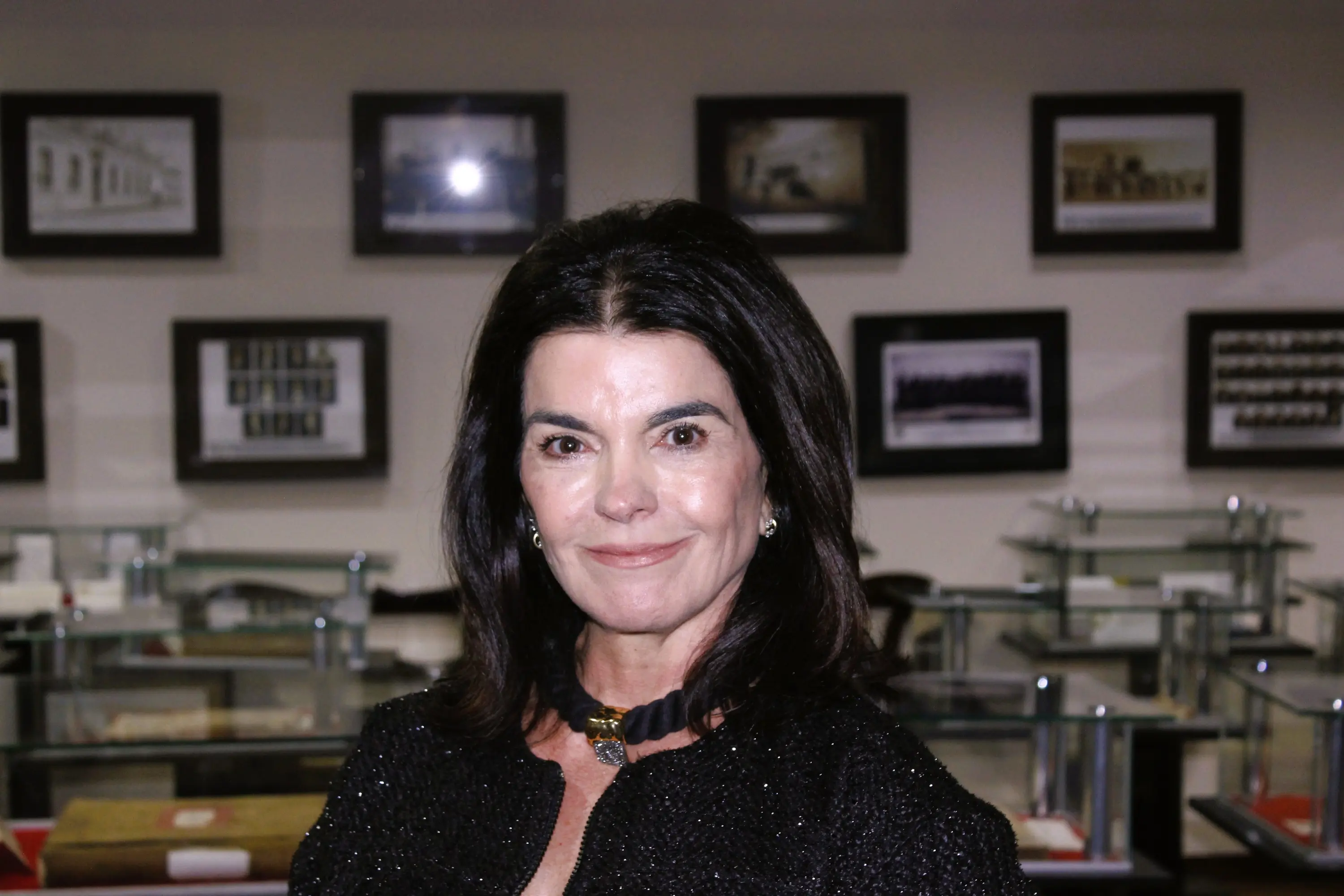 Mulher de cabelos pretos, roupa e colar pretos sorri para a câmera. Ao fundo, quadros emoldurados com fotos históricas em exposição.