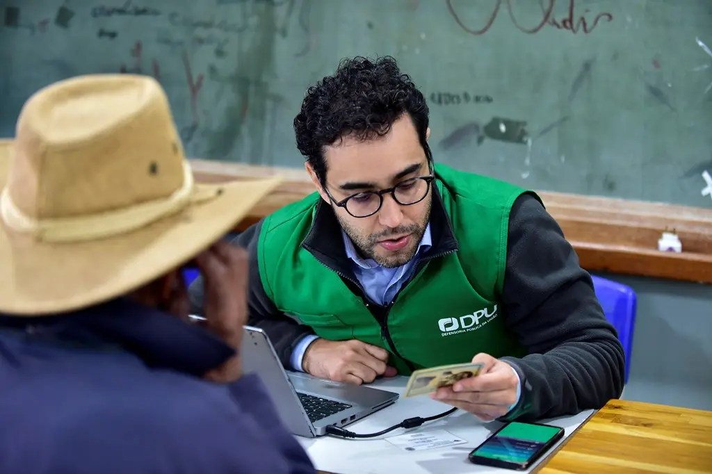 Foto que mostra um defensor público da União, vestido com colete verde da instituição, atendendo um senhor, que aparece de costas, usando chapéu e jaqueta azul marinho.