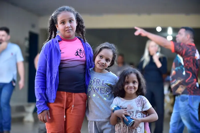 Três meninas sorriem em frente à Escola Municipal Dona Lila Hill de Souza, no distrito de Vila Picada, em Porto Esperidião-MT. A mais nova segura um copo azul nas mãos.