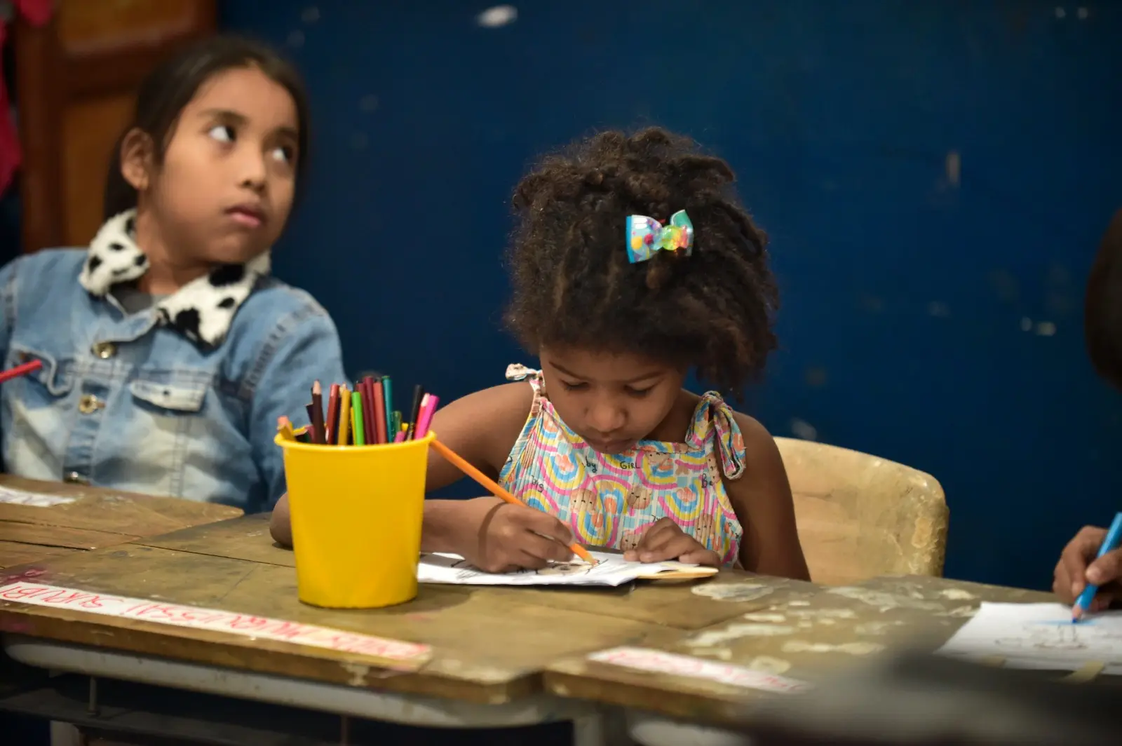 Duas meninas estão sentadas em carteira escolar. Uma delas desenha com concentração, cercada por lápis de cor em um pote amarelo. A outra observa ao lado, com expressão curiosa.