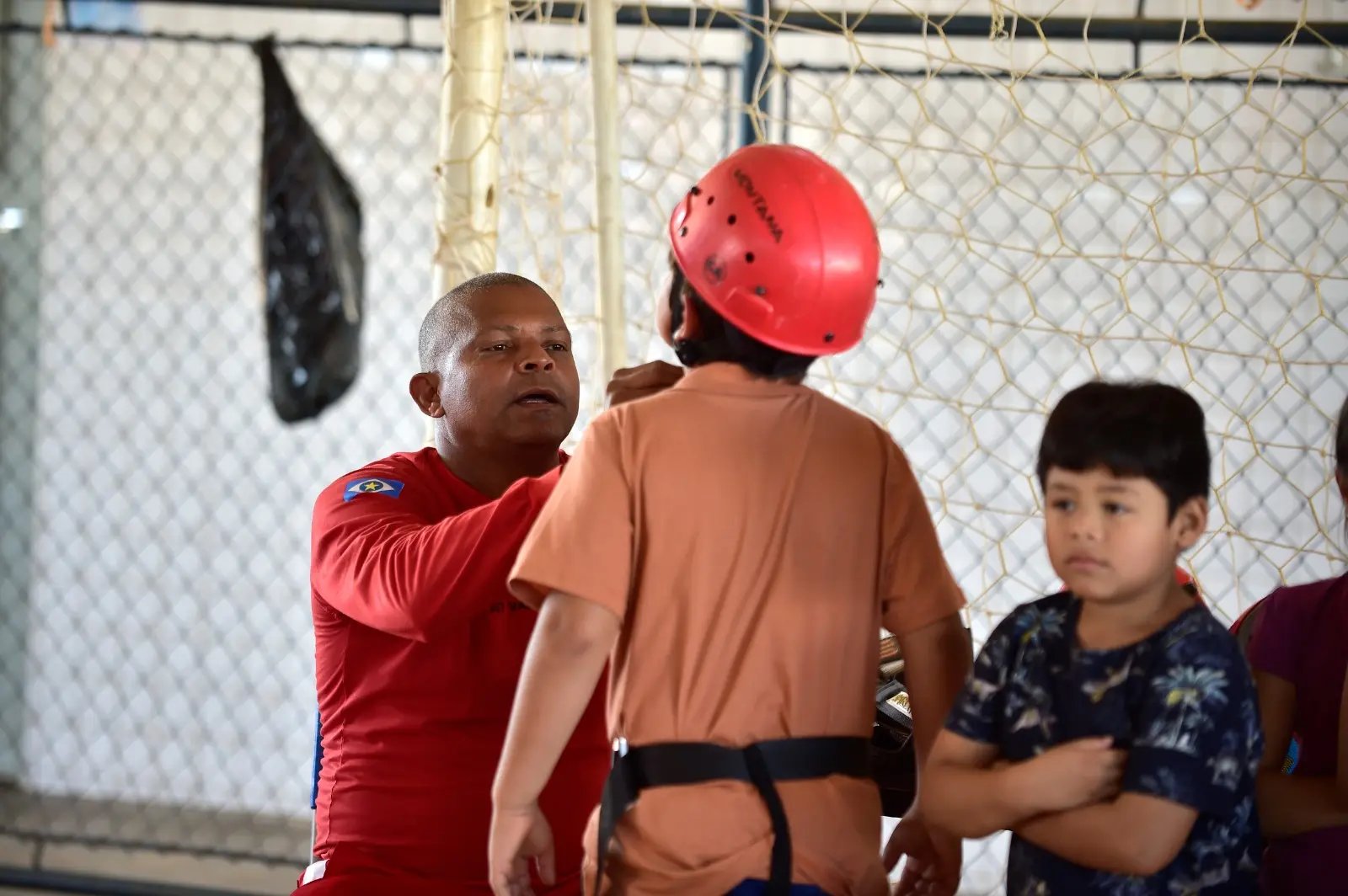 Bombeiro com uniforme vermelho ajusta o capacete de segurança de uma criança antes de atividade. Ao lado, outra criança observa. Ao fundo, rede e ambiente esportivo escolar.