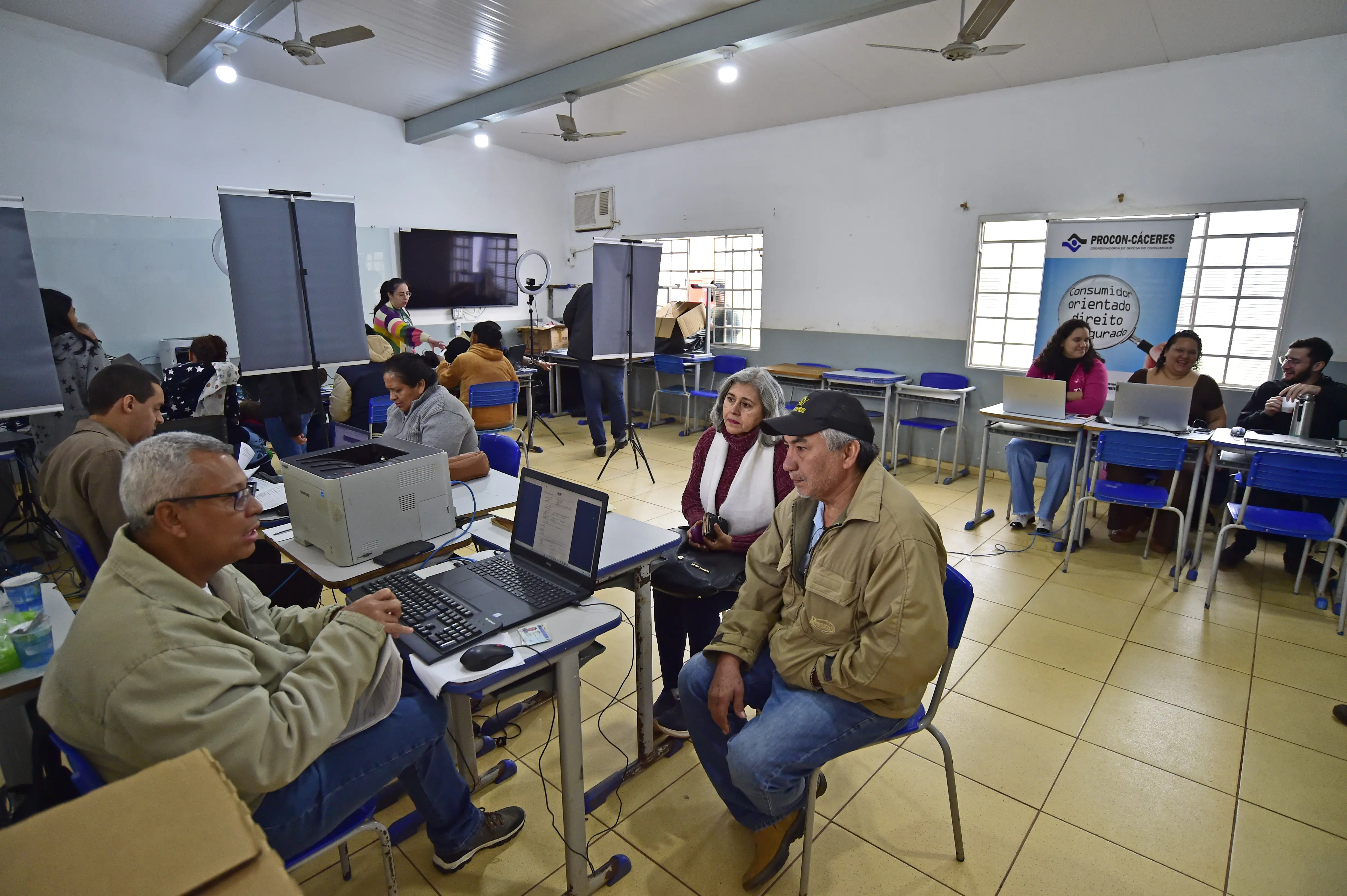 Sala de aula transformada em espaço de atendimento comunitário. Diversas mesas com computadores são utilizadas para orientar e registrar cidadãos. No centro, um casal é atendido por um servidor. Ao fundo, banner do Procon-Cáceres e equipe prestando atendimento. 