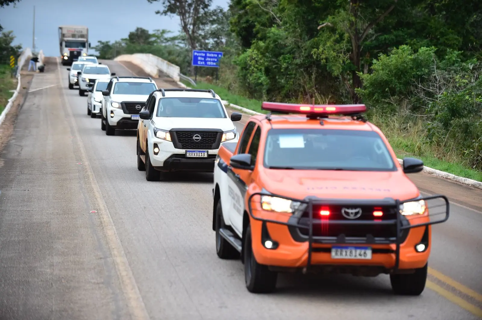 Comboio de veículos, liderado por caminhonete laranja com luzes acesas da Defesa Civil, atravessa ponte sobre o Rio Jauru. Caminhonetes brancas seguem enfileiradas sob céu nublado