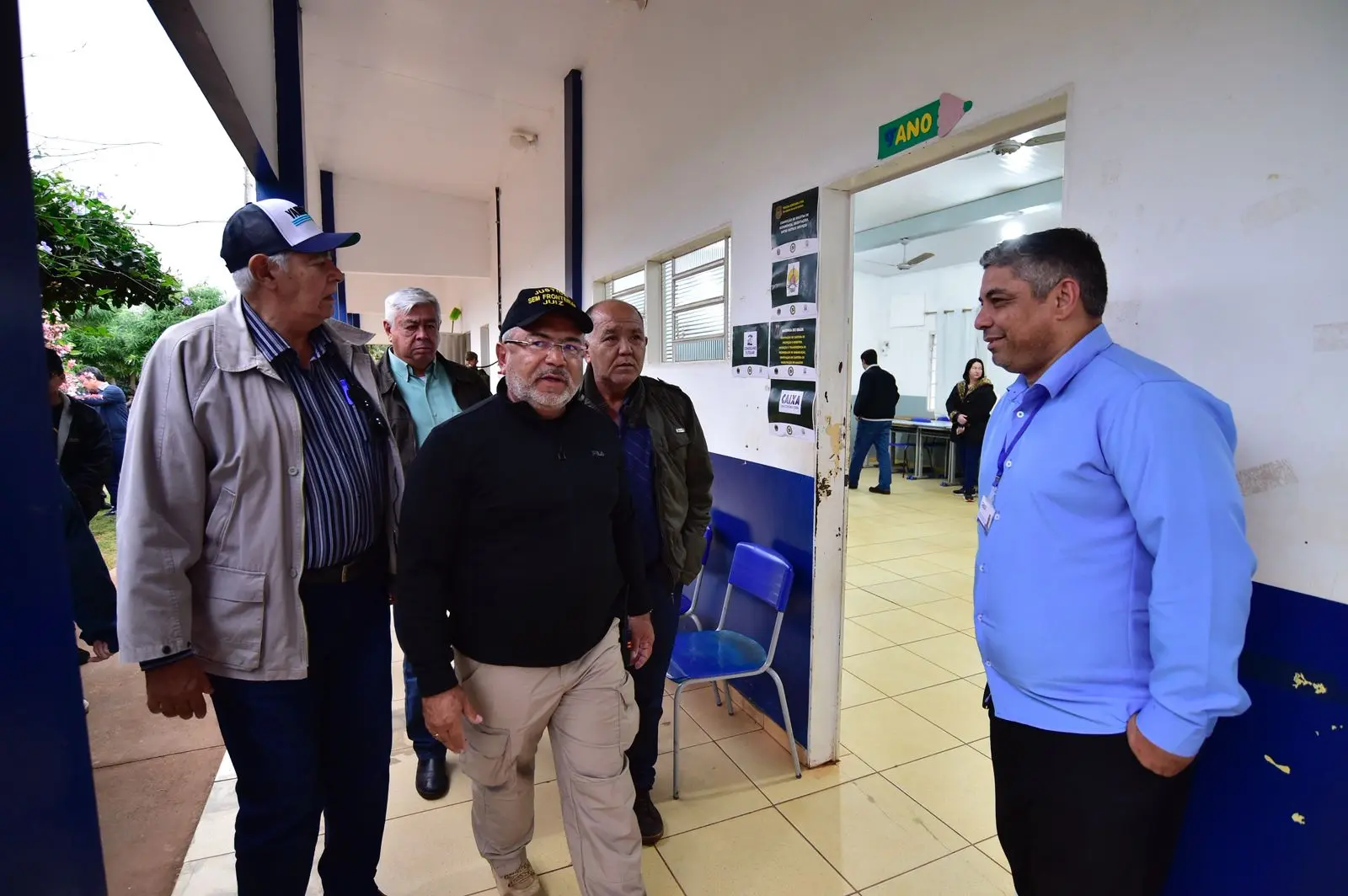 Homens caminham por corredor escolar. À direita, homem de camisa azul sorri próximo à porta da sala de aula. No fundo, ambiente interno com pessoas em atendimento e placas informativas na parede.