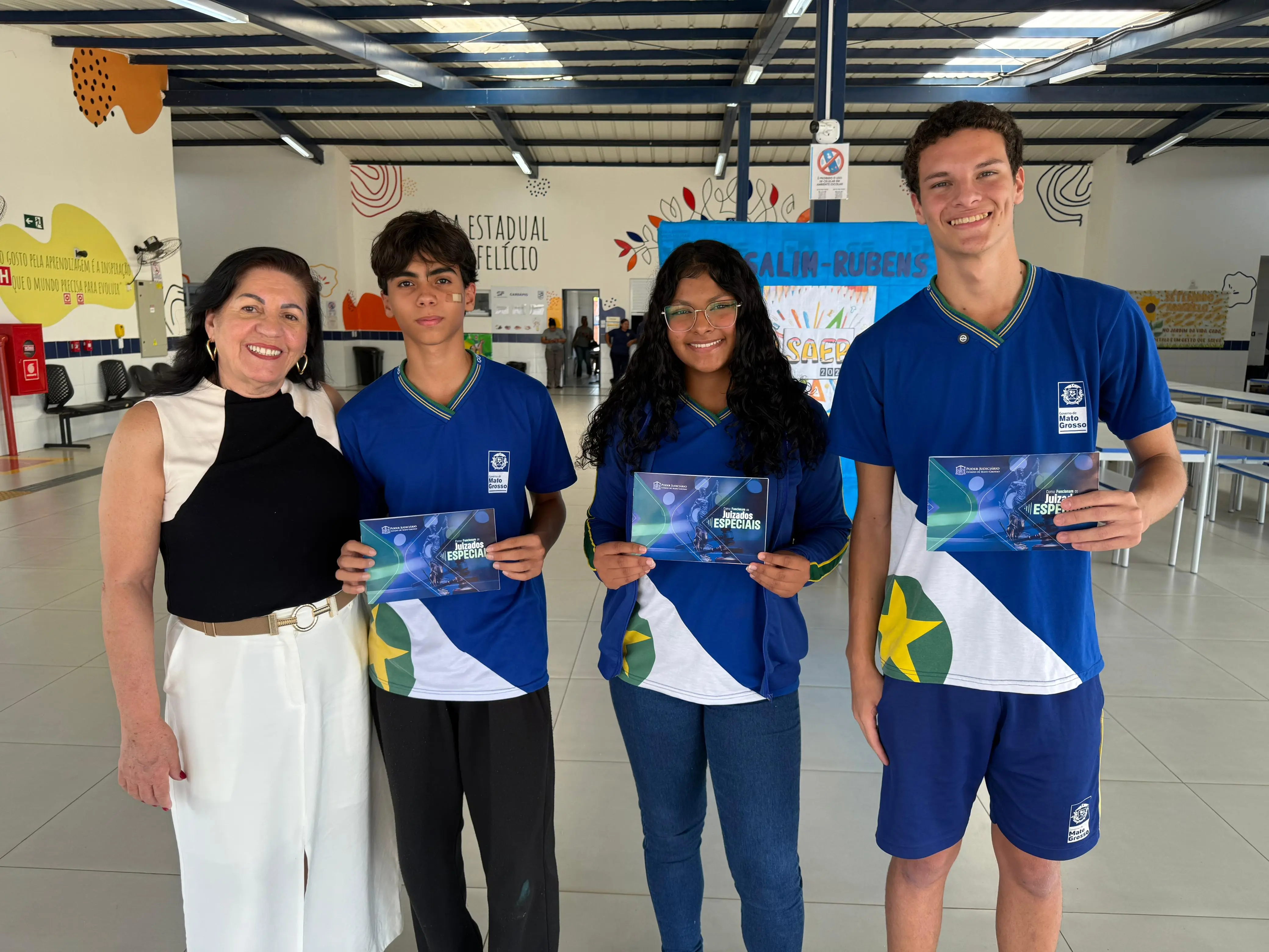 Uma mulher sorridente em preto e branco posa entre três estudantes adolescentes, dois meninos e uma menina, todos vestindo camisetas azuis com a bandeira do Brasil. Cada um segura um folheto, e eles estão em um ambiente interno e iluminado de escola