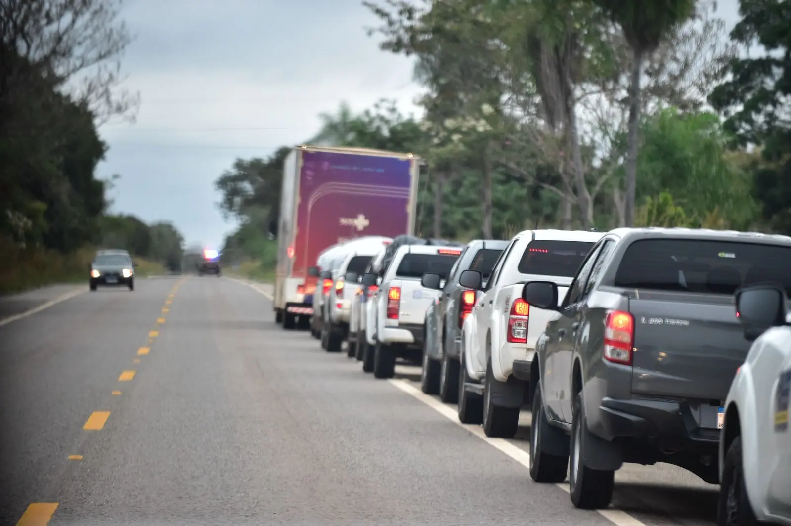 Longa fila de caminhonetes brancas e um caminhão roxo trafega por rodovia cercada por árvores. Céu cinza e luzes traseiras acesas indicam comboio em deslocamento. Viatura aparece ao fundo.