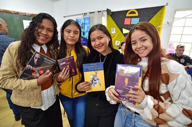 Quatro adolescentes seguram livros e sorriem para a câmera durante ação de incentivo à leitura. Atrás, cartazes coloridos decoram a sala e outras pessoas participam da atividade