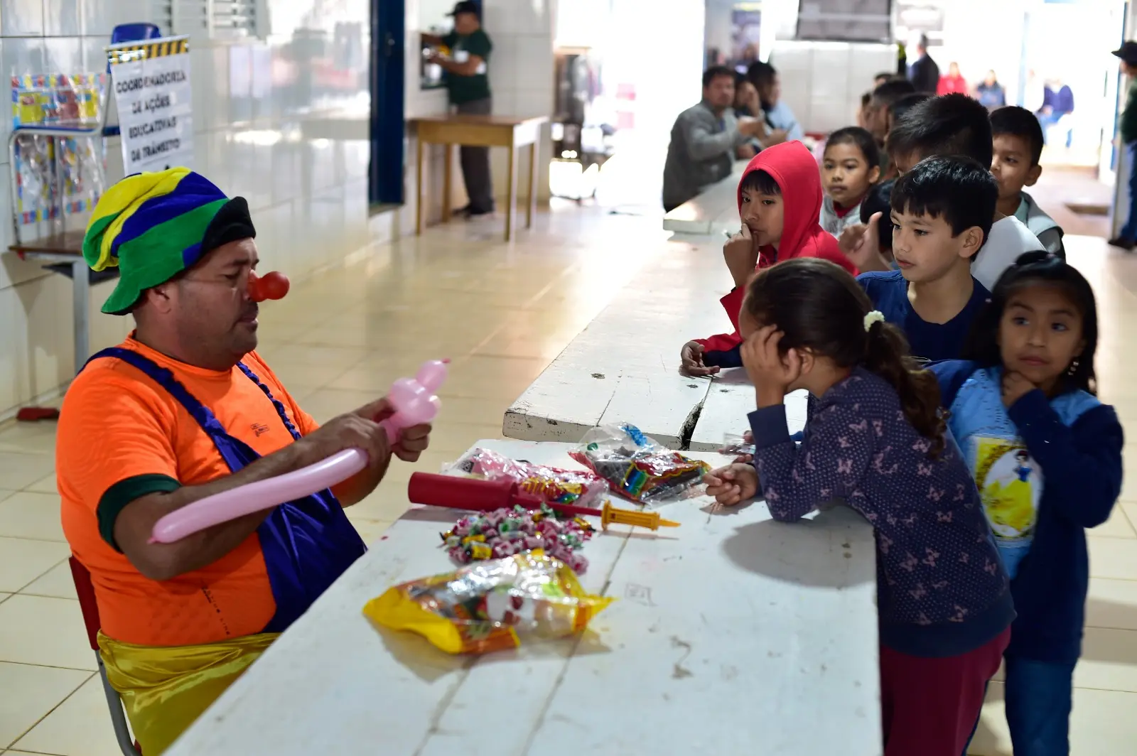 Palhaço sentado modela balões em formato de brinquedo enquanto crianças observam com atenção. Mesa tem sacos de doces e itens coloridos. Cena ocorre em espaço coberto