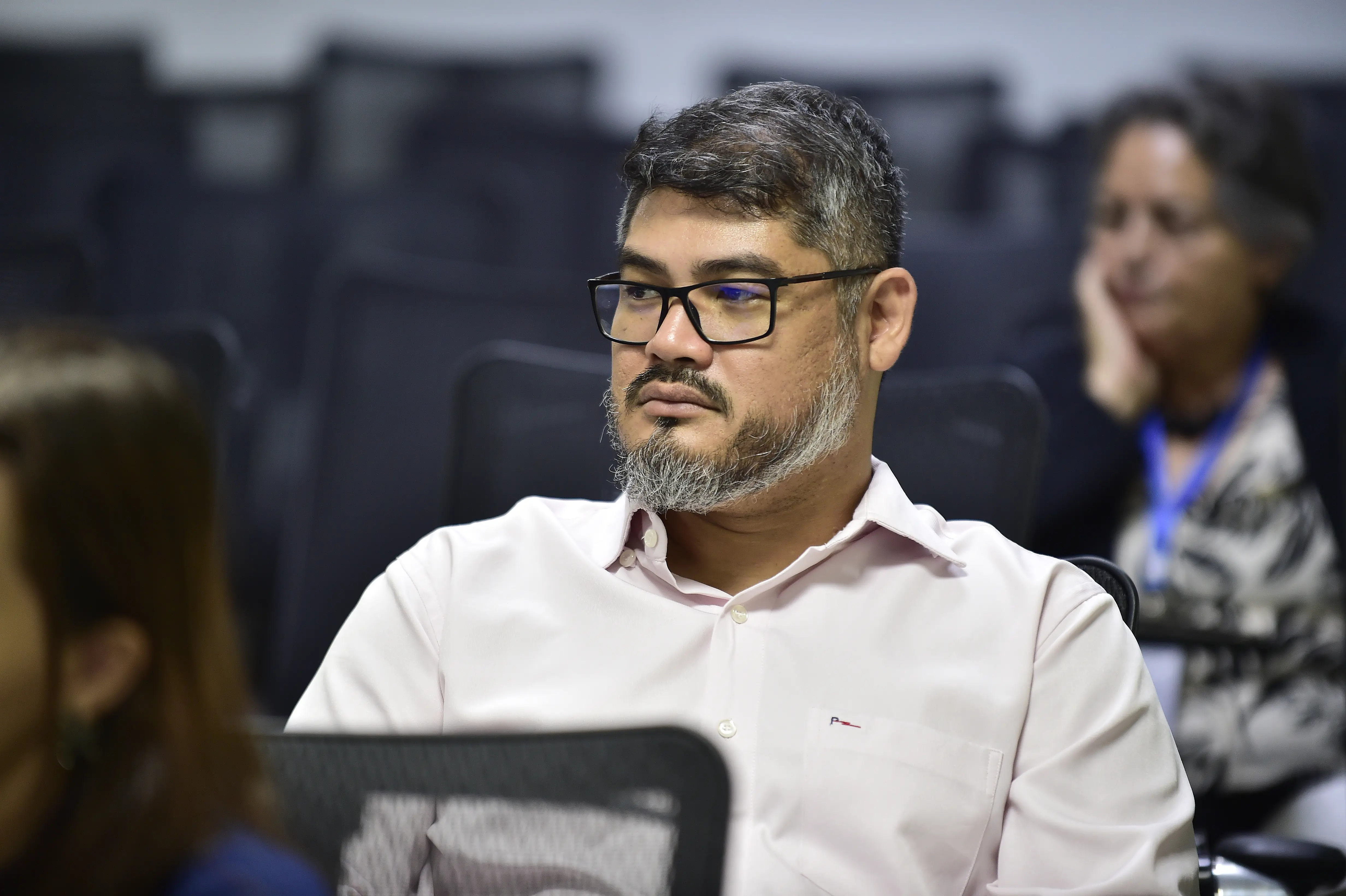 Psicólogo Dyumdy Araújo sentado em meio aos demais participantes do curso. Ele é um homem negro, de olhos puxados, cabelos lisos e grisalhos, usando óculos de grau de armação preta e camisa social rosa clara. 