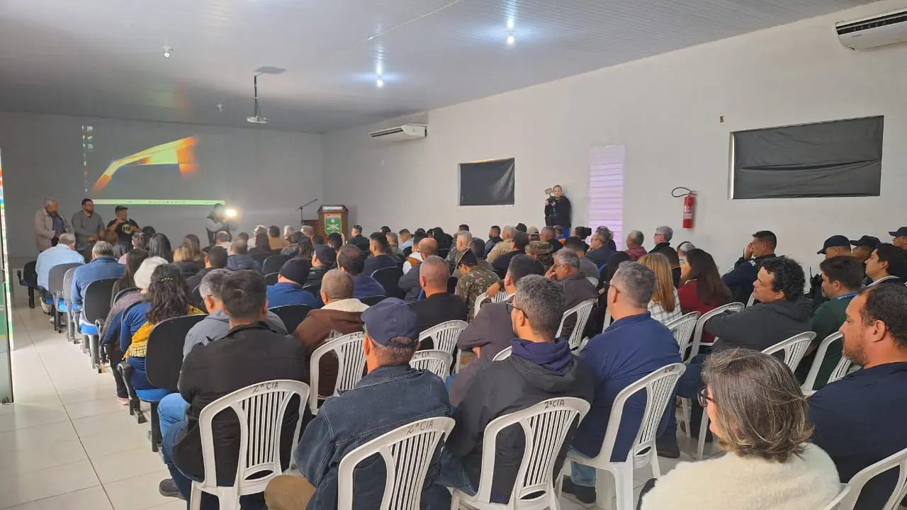 Auditório cheio, com homens e mulheres sentados em cadeiras brancas e azuis, assistindo à apresentação. Na frente, pessoas em pé falam ao microfone. Tela de projeção exibe imagem no fundo