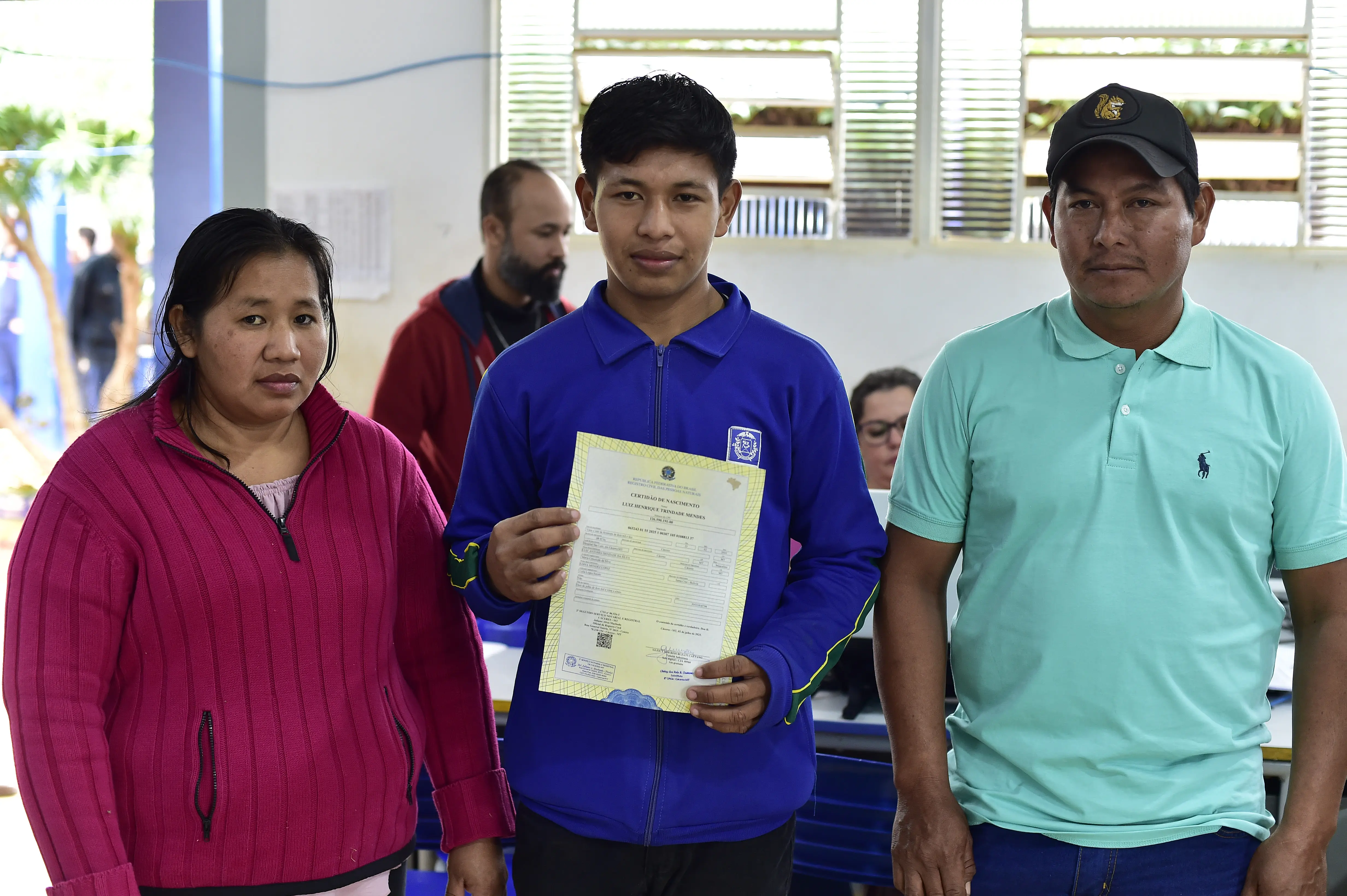 Em ambiente interno, jovem veste jaqueta azul e segura uma certidão de nascimento com as duas mãos. Ao seu lado, estão uma mulher de blusa rosa e um homem de boné e camiseta verde. Todos encaram a câmera com expressões serenas.