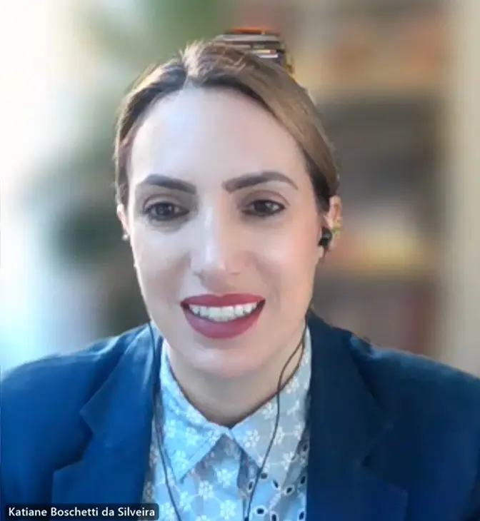 Close da participante Katiane Boschetti da Silveira durante a videoconferência. Ela usa blazer azul e camisa clara, fones de ouvido e aparece sorridente, em ambiente iluminado, transmitindo profissionalismo.