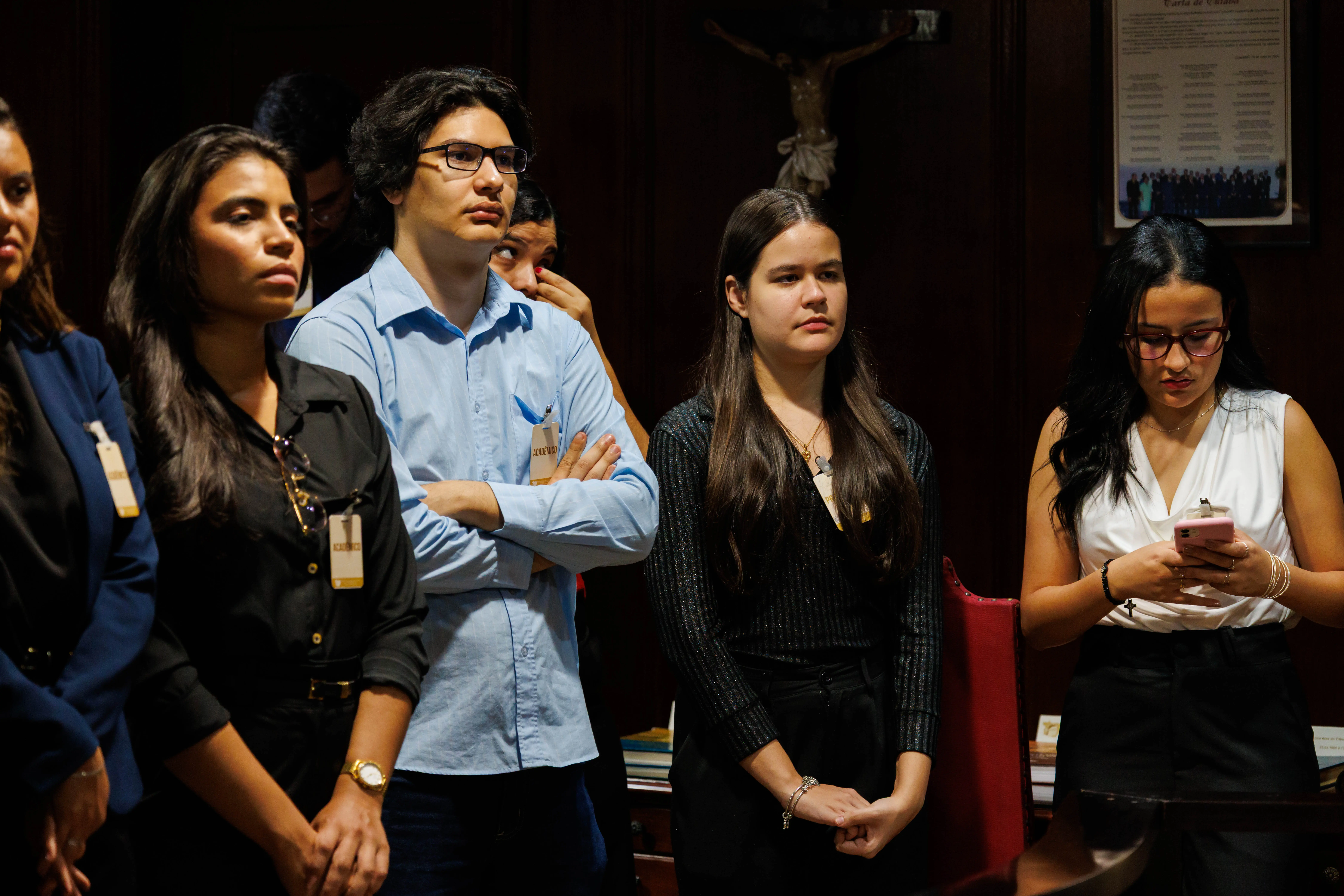 Grupo de jovens observa explicações no Espaço Memória. Alguns usam crachás de acadêmicos, enquanto uma participante manuseia celular. Ao fundo, crucifixo e quadro reforçam o tom formal do espaço.