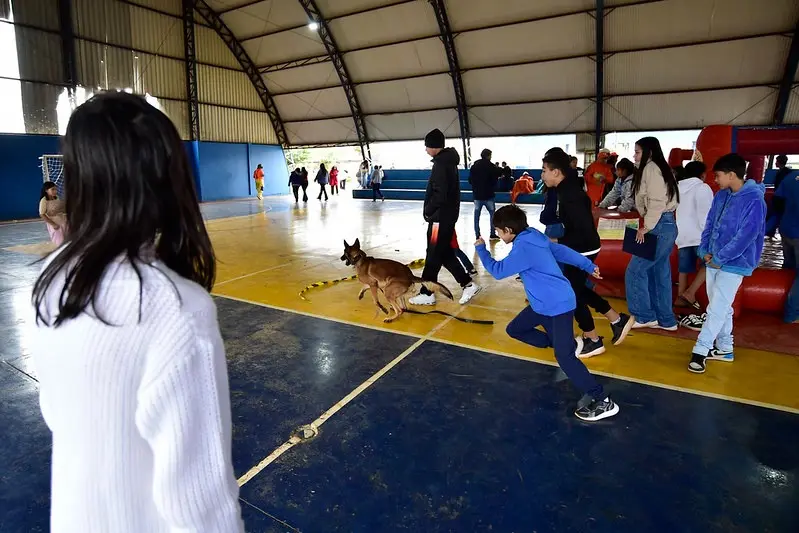 Crianças correm e interagem com um cão policial dentro de uma quadra coberta. Outras observam ao redor, criando um ambiente dinâmico e animado de convivência e integração.