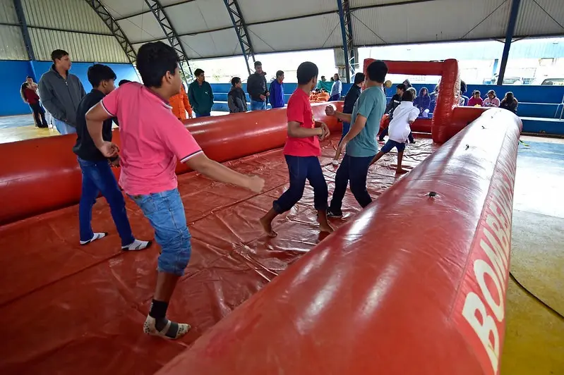 Crianças e adolescentes brincam em um campo inflável de futebol de sabão, com piso vermelho escorregadio e bordas altas. A atividade ocorre em ginásio coberto e conta com público assistindo ao fundo. Perguntar ao ChatGPT