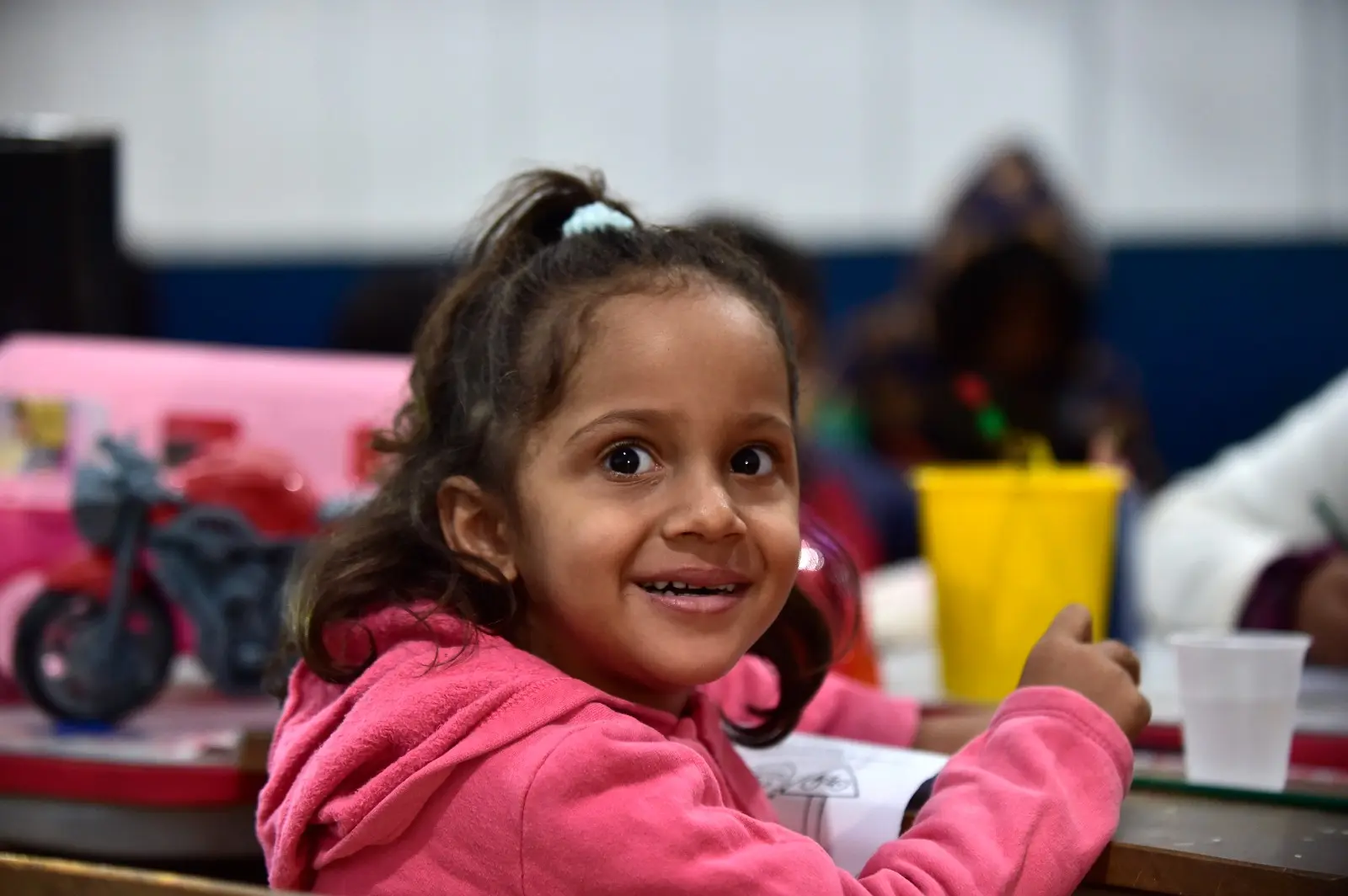Menina com moletom rosa sorri para a câmera enquanto desenha na sala. Atrás dela, colegas também participam da atividade. Há brinquedos sobre a mesa e ambiente alegre.