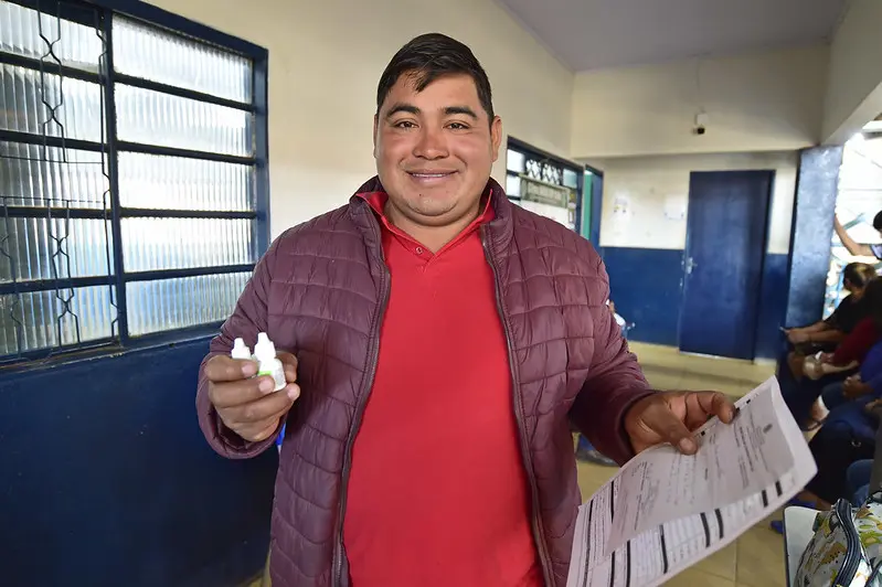 Foto que mostra um homem segurando remédios e receita médica nas mãos. Ele está em pé o corredor de uma escola e sorrindo para a foto. Homem negro, com traços bolivianos (cabelos lisos e pretos e olhos puxados), usando camiseta vermelha e jaqueta marsala.
