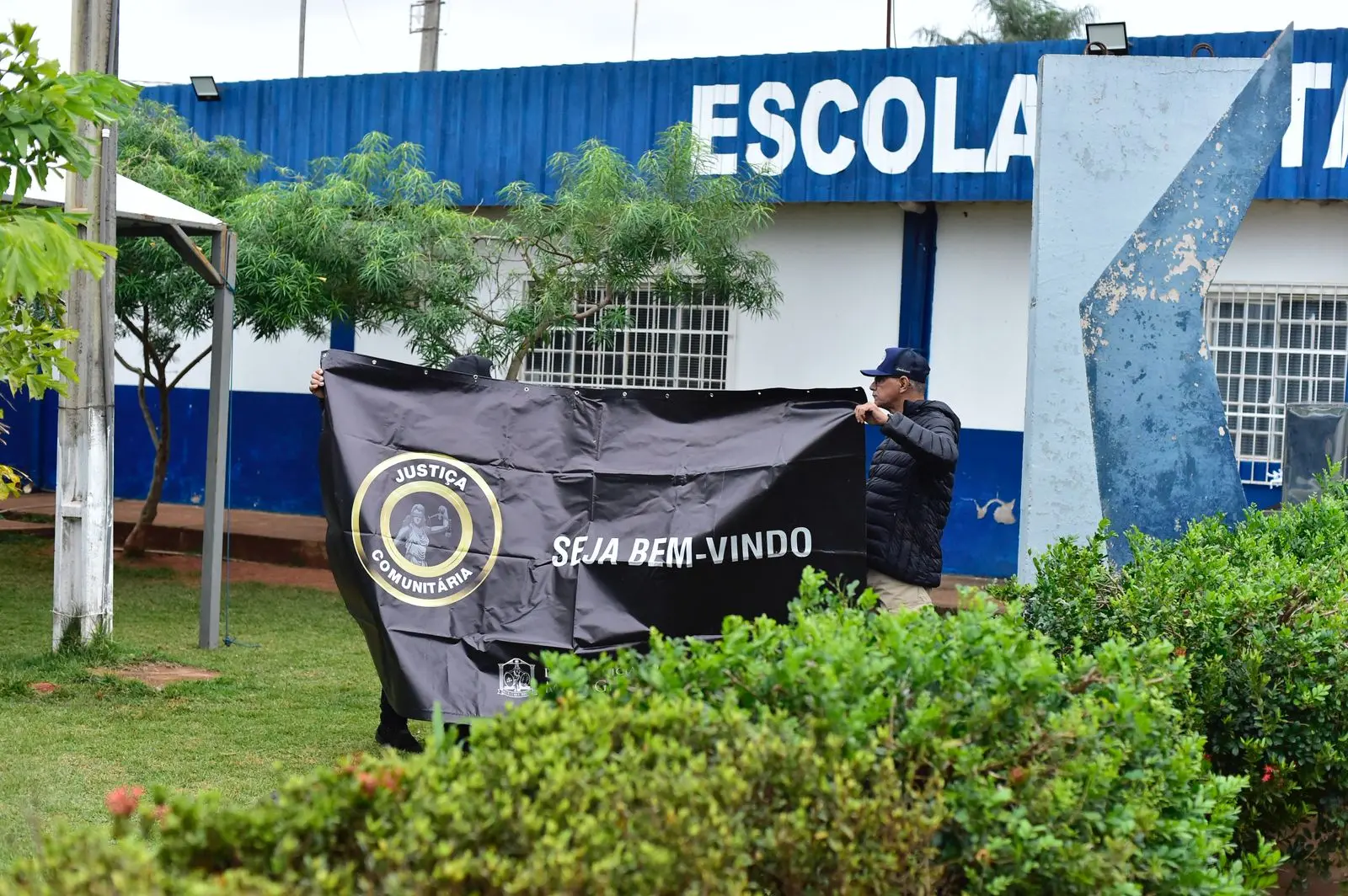 Dois homens seguram faixa preta com os dizeres "Justiça Comunitária – Seja Bem-vindo", em frente à fachada da Escola 12 de Outubro. Há arbustos e árvores no jardim e parte do letreiro da escola ao fundo